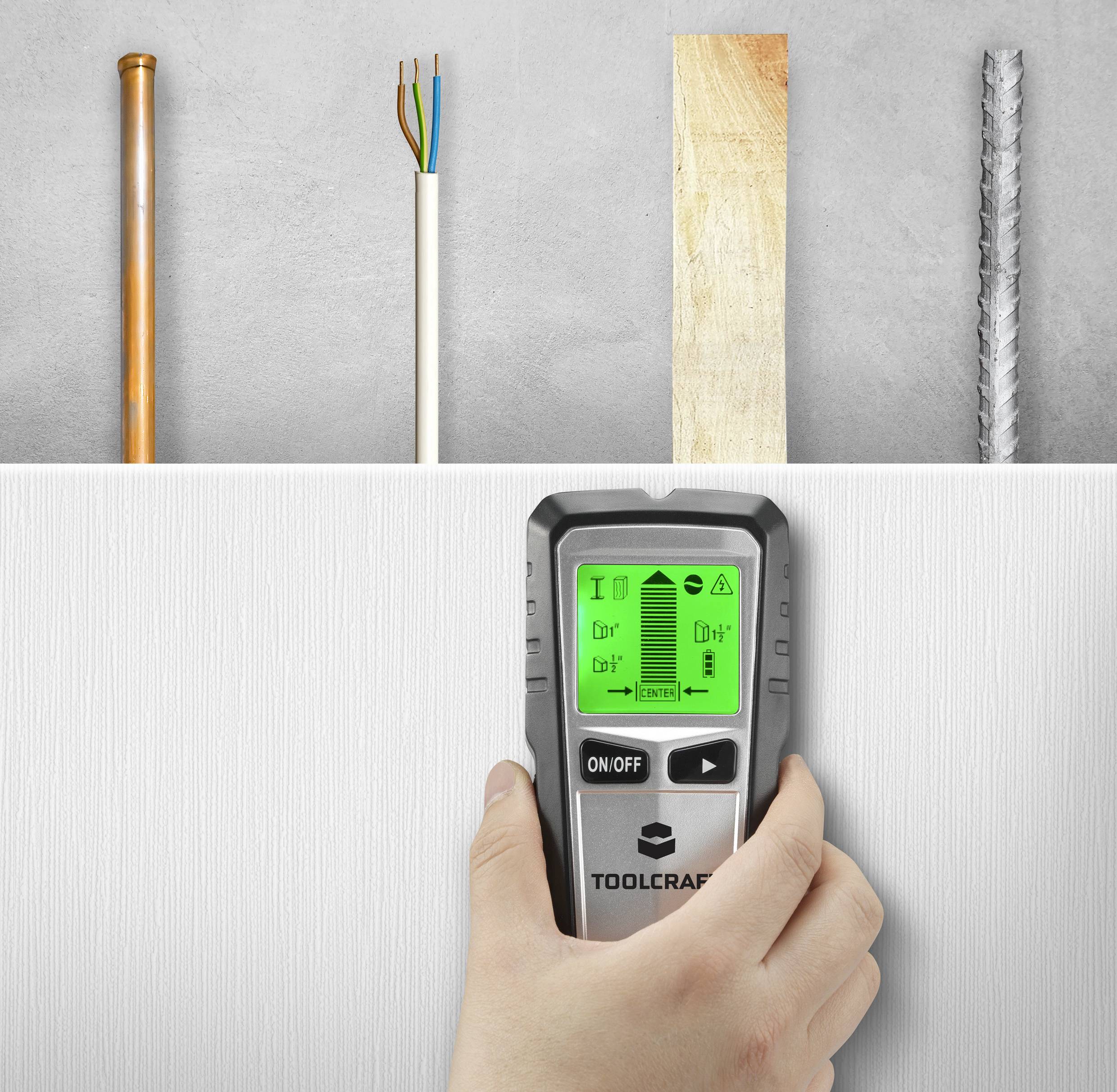 Eine Hand hält einen Detektor über eine weiße Wand. Der Bildschirm zeigt Symbole für Metall, Holz und elektrische Leitungen an. Im Hintergrund sind Rohre, Kabel, ein Holzbrett und eine Metallstange zu sehen.