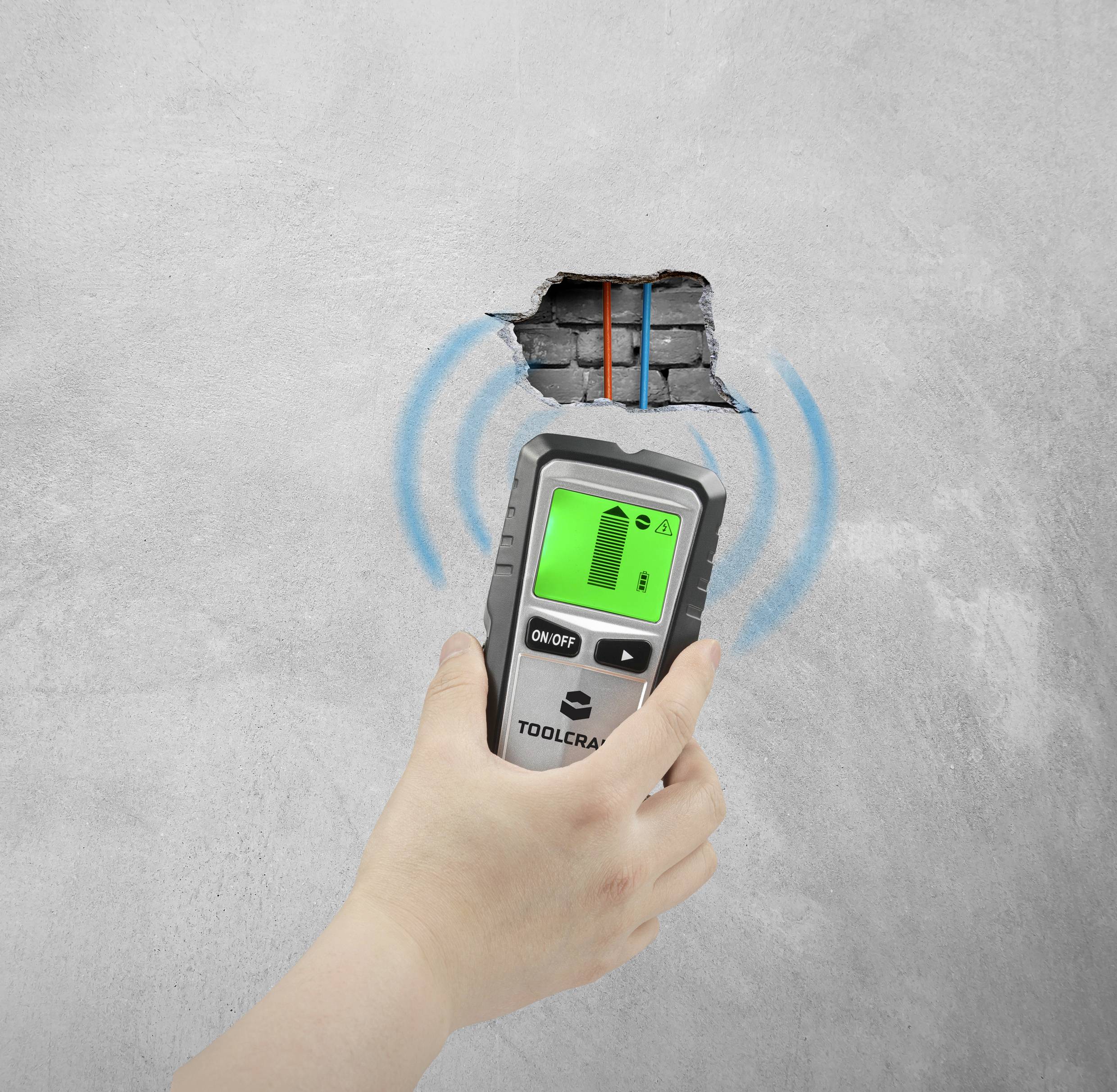 Eine Person hält ein Gerät zur Wandsuche, das Leitungen hinter der Betonwand aufspürt. Ein Bildschirm zeigt die Detektion an.