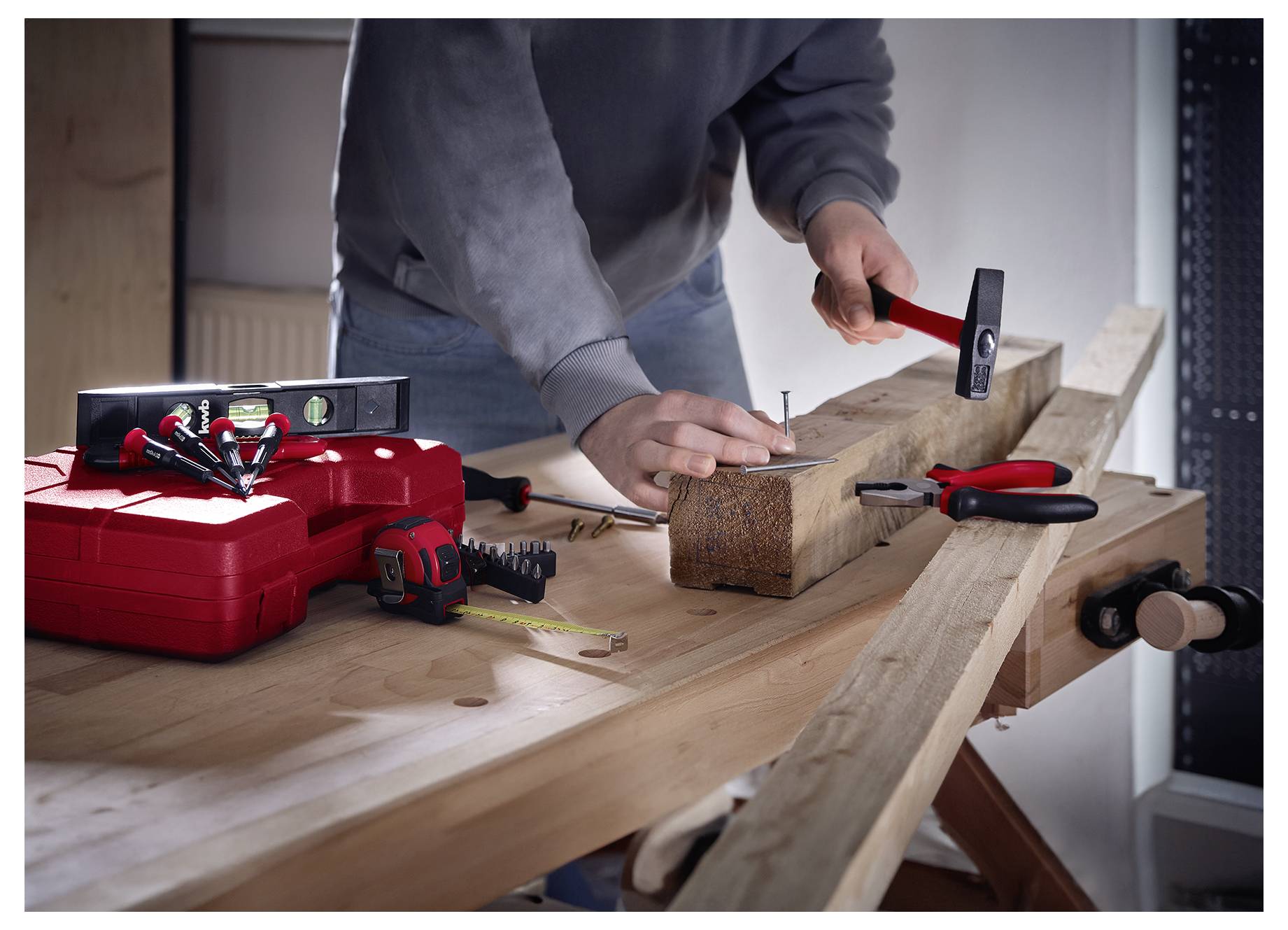 Eine Person mit einem grauen Sweatshirt hämmert einen Nagel in einen Holzbalken auf einer Werkbank. Eine rote Werkzeugbox und Werkzeuge befinden sich in der Nähe.