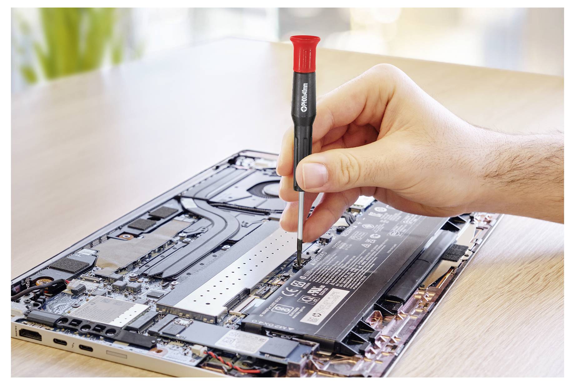 Eine Hand benutzt einen Schraubendreher, um einen Akku aus dem Inneren eines geöffneten Laptops zu entfernen und zeigt seine Komponenten auf einem Holztisch.