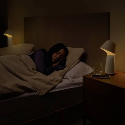 Eine Frau schläft in einem verdunkelten Schlafzimmer, eingekuschelt unter einer Decke. Auf dem Nachttisch steht ein Glas Wasser neben einer Lampe.
