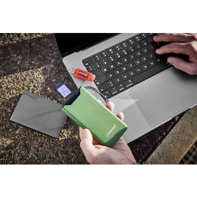 Eine Person hält eine grüne externe Festplatte vor einem Laptop. Daneben liegen ein USB-Stick und eine blaue SD-Karte auf einem Tisch.
