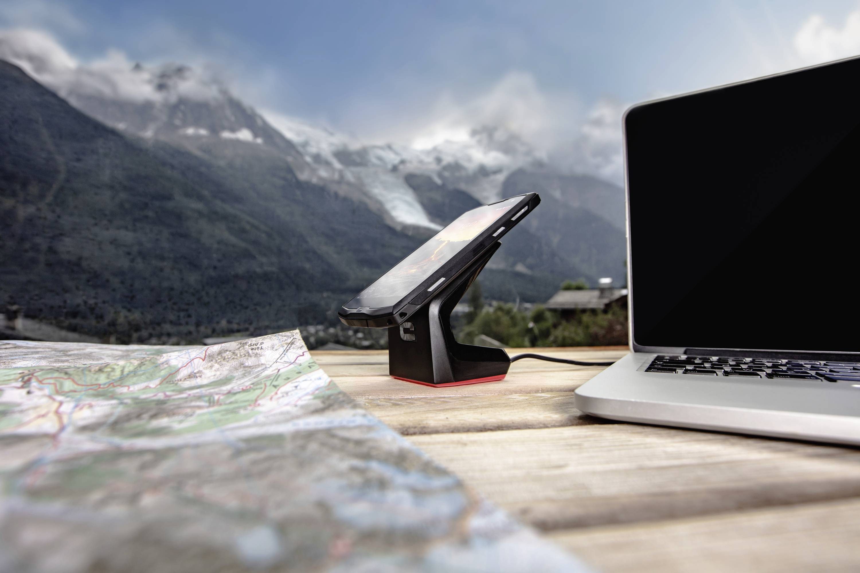Ein Laptop und ein aufgeladenes Smartphone stehen auf einem Tisch im Freien. Im Vordergrund liegt eine Landkarte, im Hintergrund Berge.