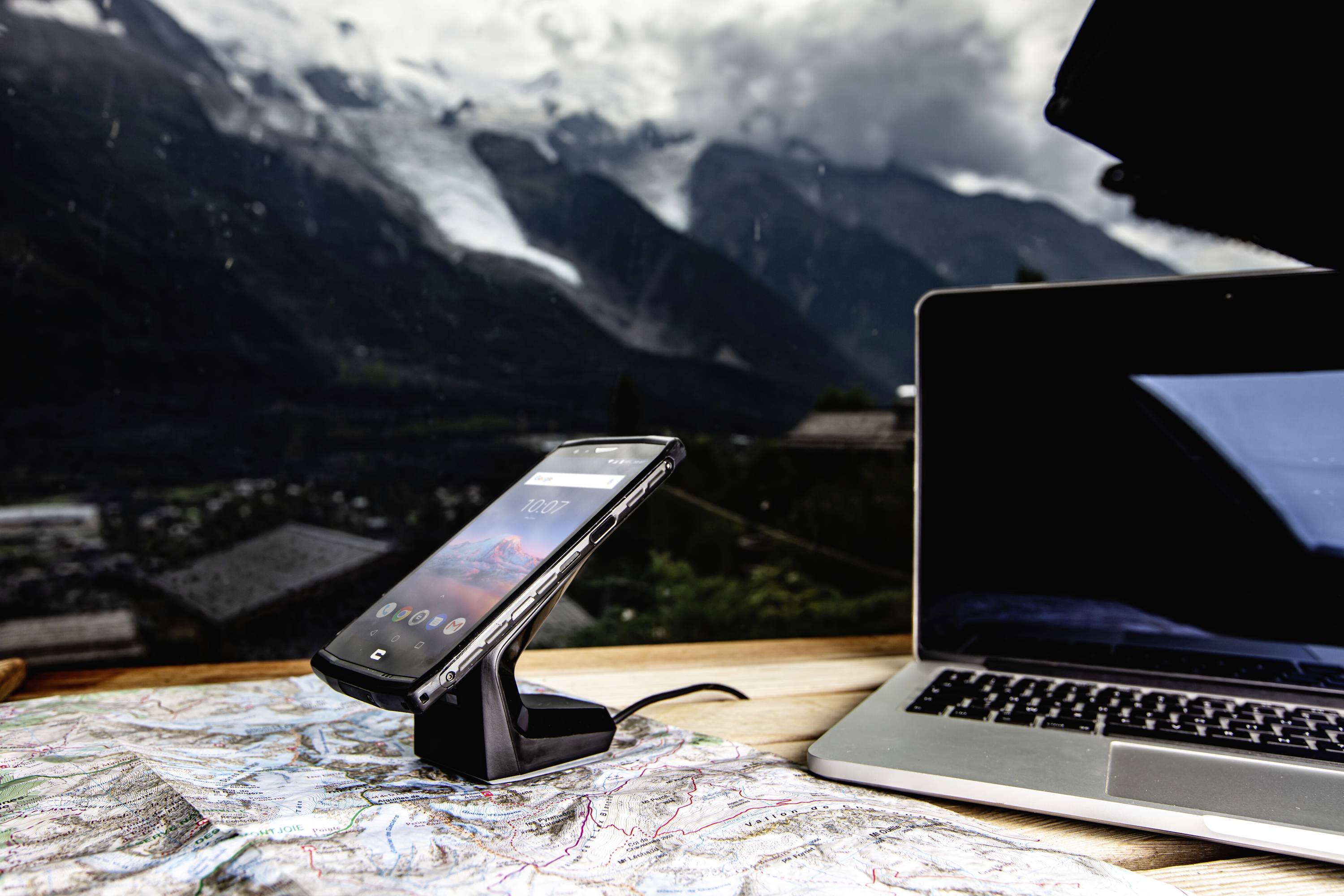 Smartphone auf Ladestation und Laptop auf Landkarte vor Berglandschaft. Fenster spiegelt Wolken und Berge.
