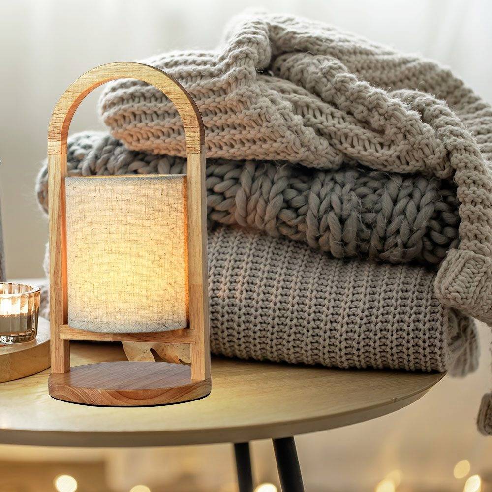 Ein runder Tisch mit einer dekorativen Lampe aus Holz und Stoff, daneben gefaltete Strickdecken in ruhigen Tönen, die ein gemütliches Ambiente schaffen.