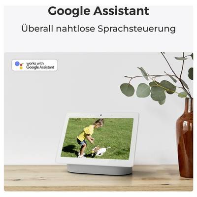 Ein Smart Display auf einer Holzoberfläche zeigt ein Kind, das mit einem Hund auf einem Feld spielt. Der Text lautet: 'Google Assistant: Überall nahtlose Sprachsteuerung.'