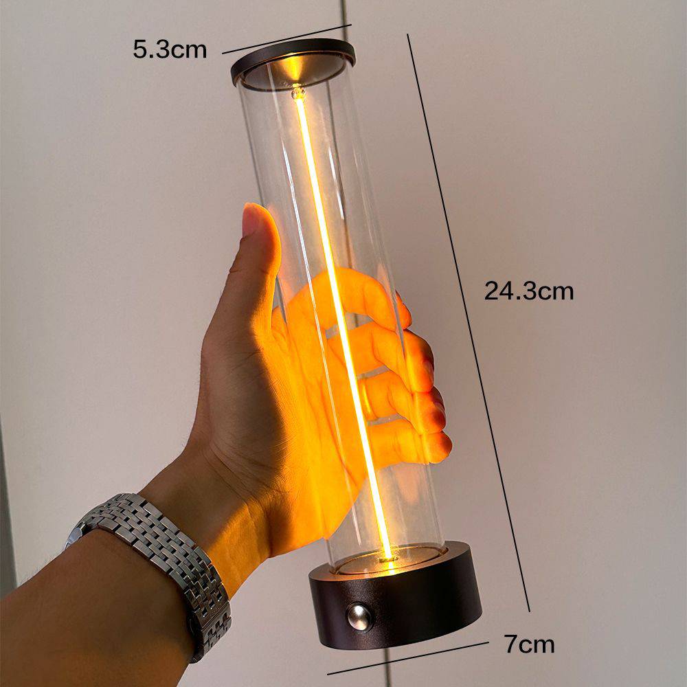 Eine Hand hält eine transparente zylindrische Lampe mit warmem Licht. Maße: 24,3 cm hoch, 5,3 cm breit, Sockel 7 cm.