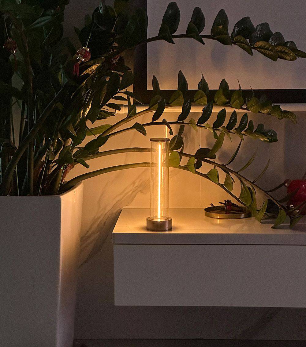 Eine beleuchtete Tischlampe mit warmem Licht steht auf einem weißen Tisch neben einer Pflanze in einem weißen Blumentopf.