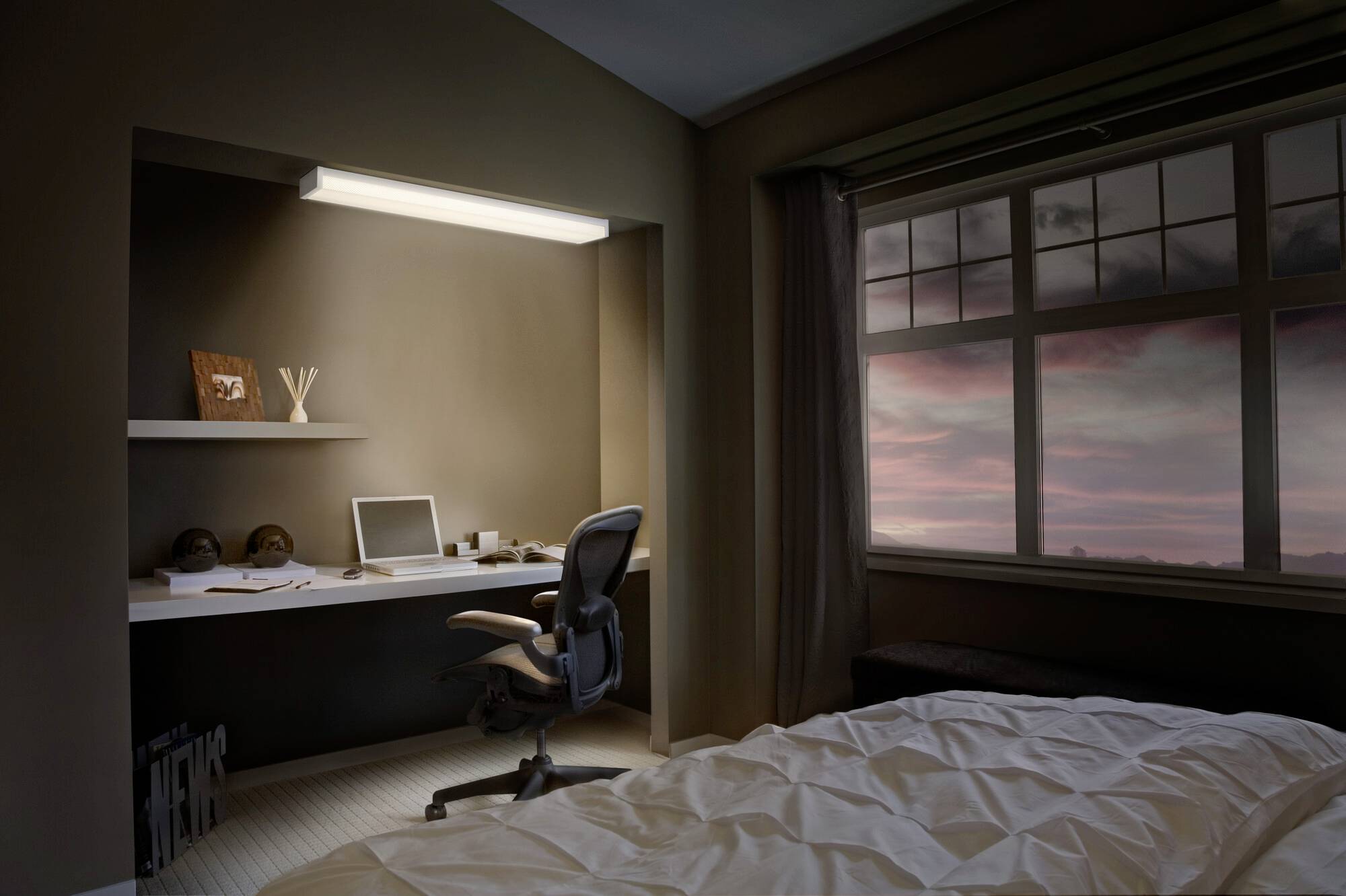 Wohnzimmer mit Schreibtisch, Computer und Stuhl, daneben ein Fenster mit Blick auf einen rosafarbenen Sonnenuntergangshimmel.