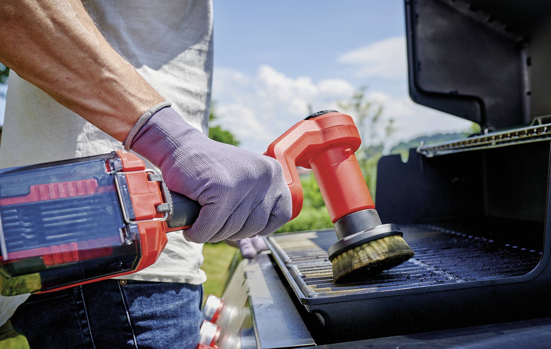 Eine Person reinigt den Grillrost eines Grills im Freien mit einer roten elektrischen Reinigungsbürste.