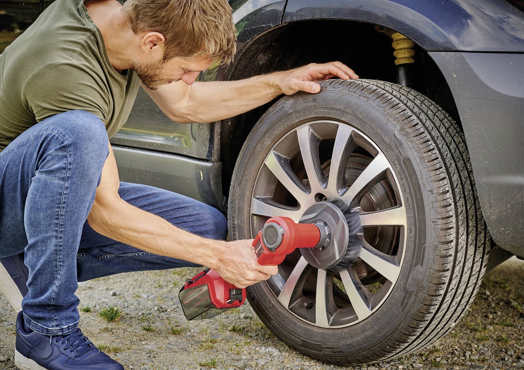 Ein Mann wechselt ein Autoreifen mit einem elektrischen Werkzeug. Er dreht die Radmuttern eines grauen Autos an einem sonnigen Tag fest.