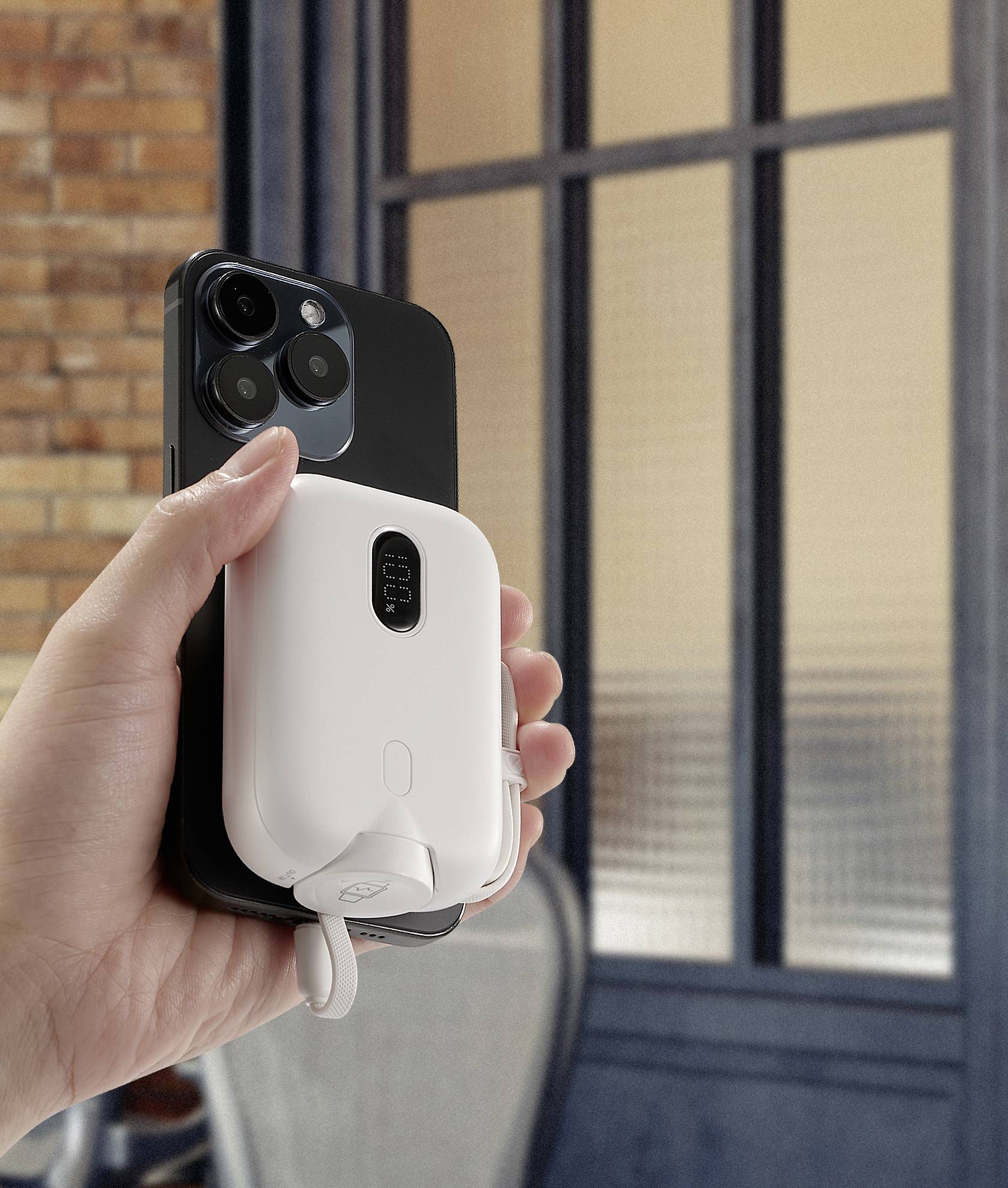 Eine Hand hält ein Smartphone mit einem weißen Akku-Pack daran befestigt. Im Hintergrund sind Fenster und Ziegelwände zu sehen.