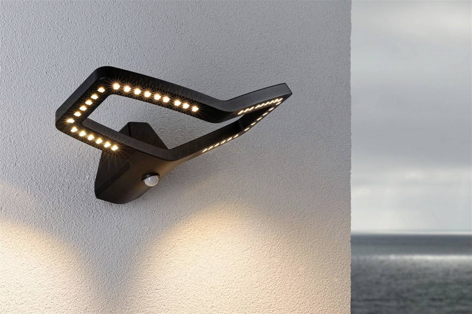 Moderne Außenwandleuchte aus schwarzem Metall mit LED-Licht, montiert an einer weißen Wand, Meer im Hintergrund.