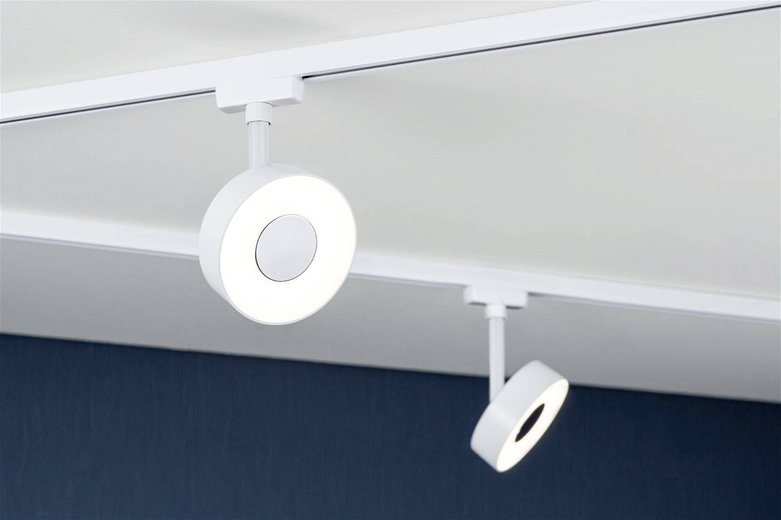 Zwei runde, weiße Deckenleuchten an einer Schiene montiert, strahlen Licht aus. Sie befinden sich in einem Raum mit dunkelblauer Wand.