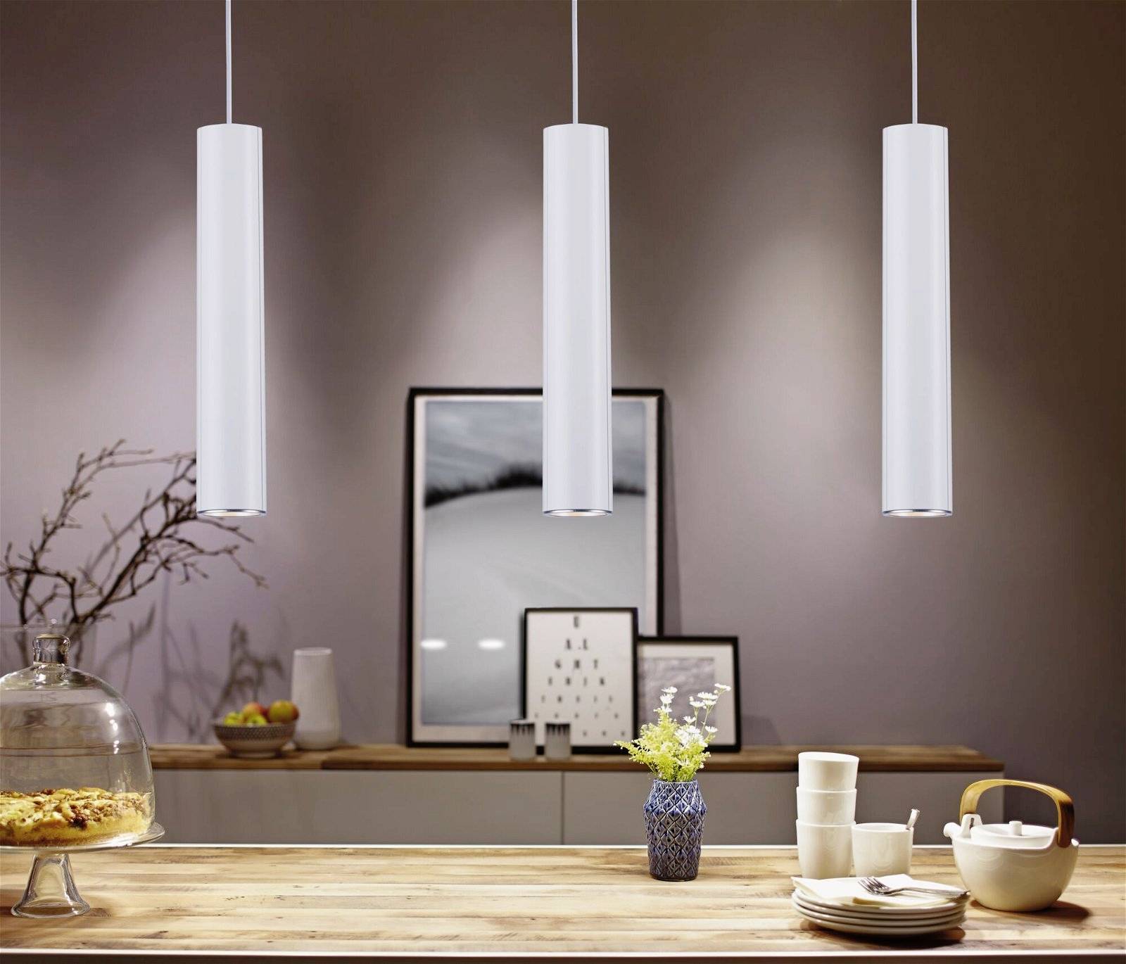 Drei weiße Pendelleuchten hängen über einem Holztisch mit Geschirr und Blumen. Im Hintergrund sind minimalistische Kunstwerke an der Wand.