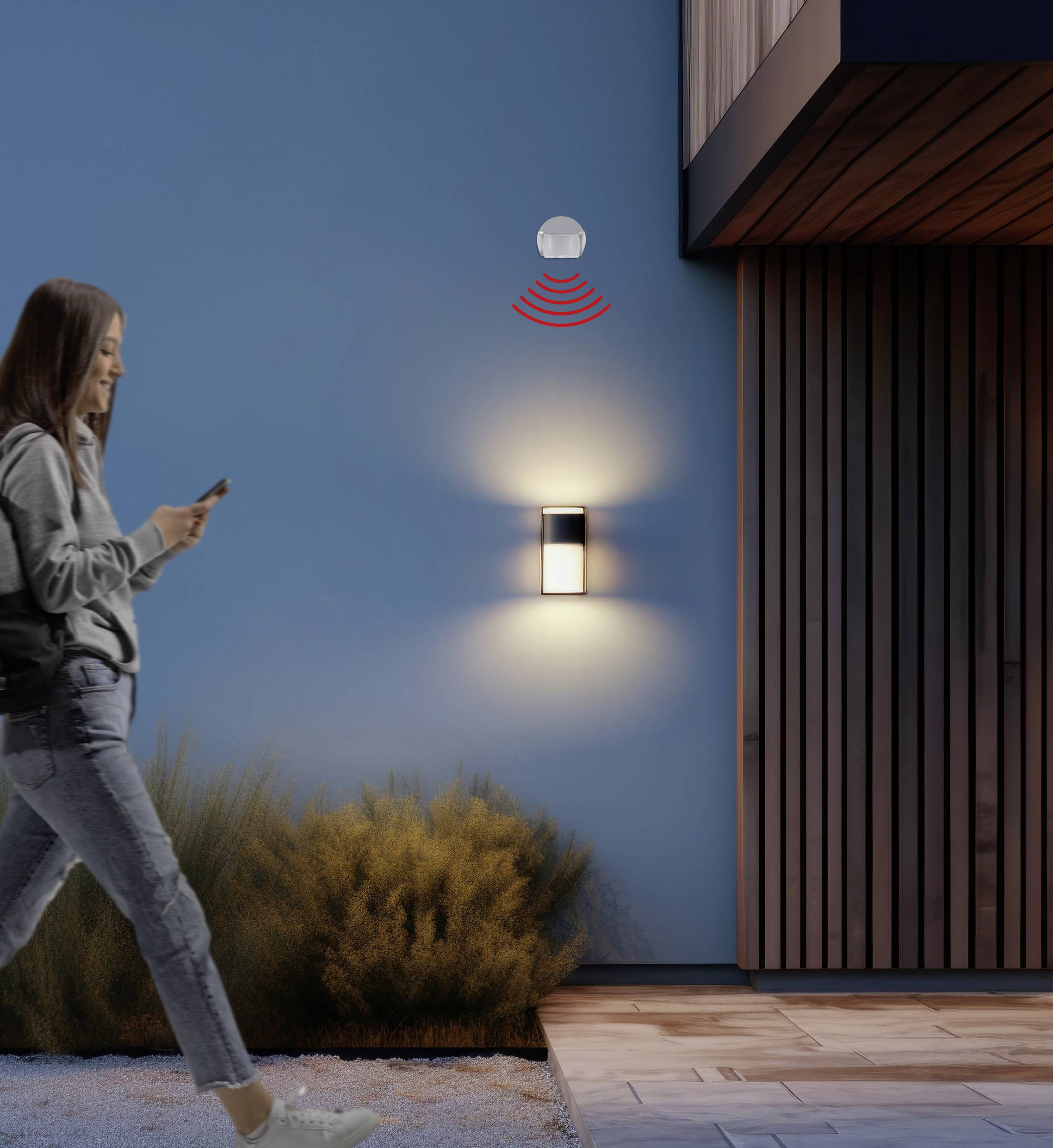 Eine Frau geht an einem beleuchteten Gebäude vorbei, während sie auf ihr Smartphone schaut. Eine Lampe und ein Bewegungssensor sind an der Wand montiert.