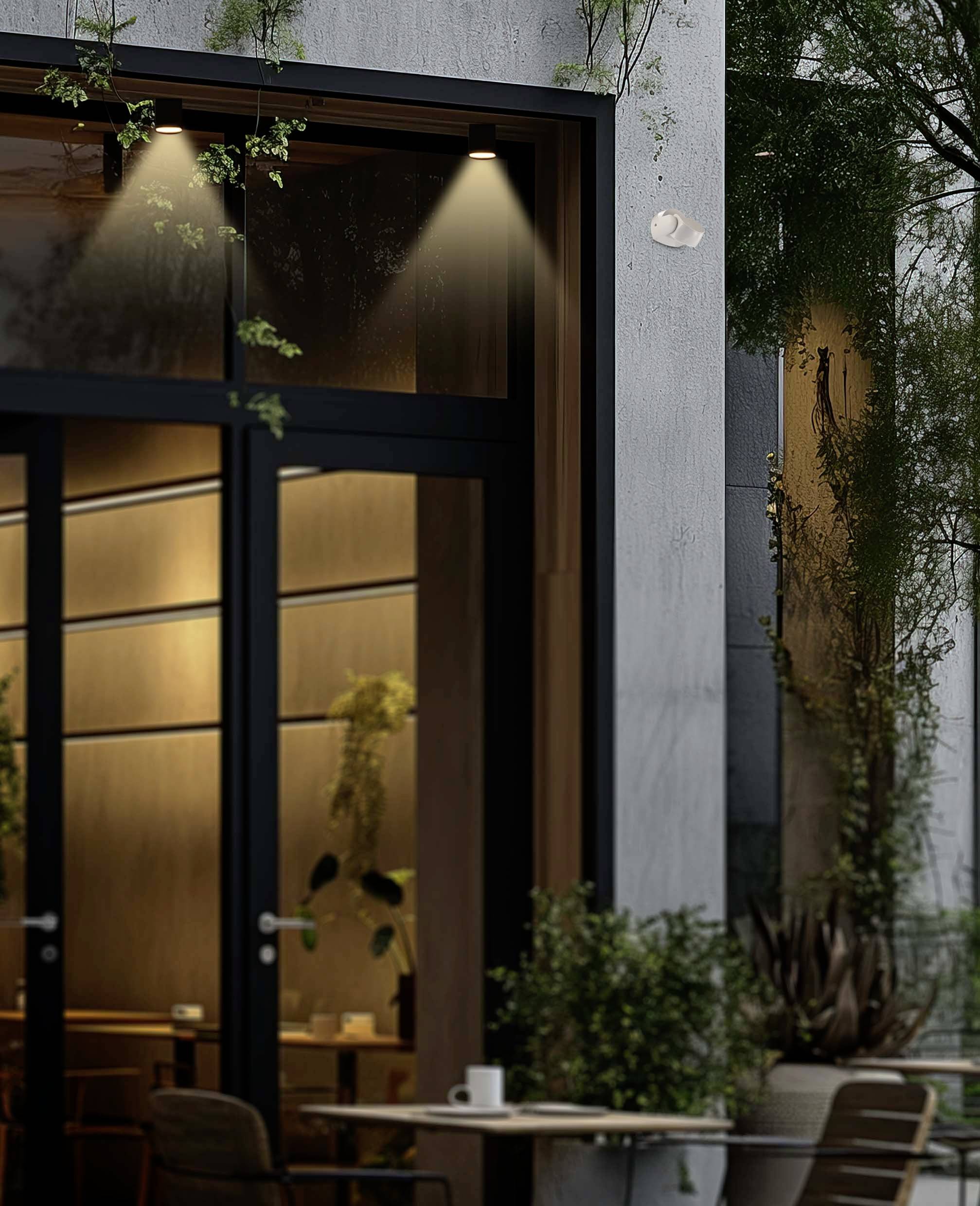 Außenseite eines modernen Cafés bei Dämmerung, warm beleuchtete Fenster, Pflanzen an der Fassade, Tisch und Stühle im Vordergrund.