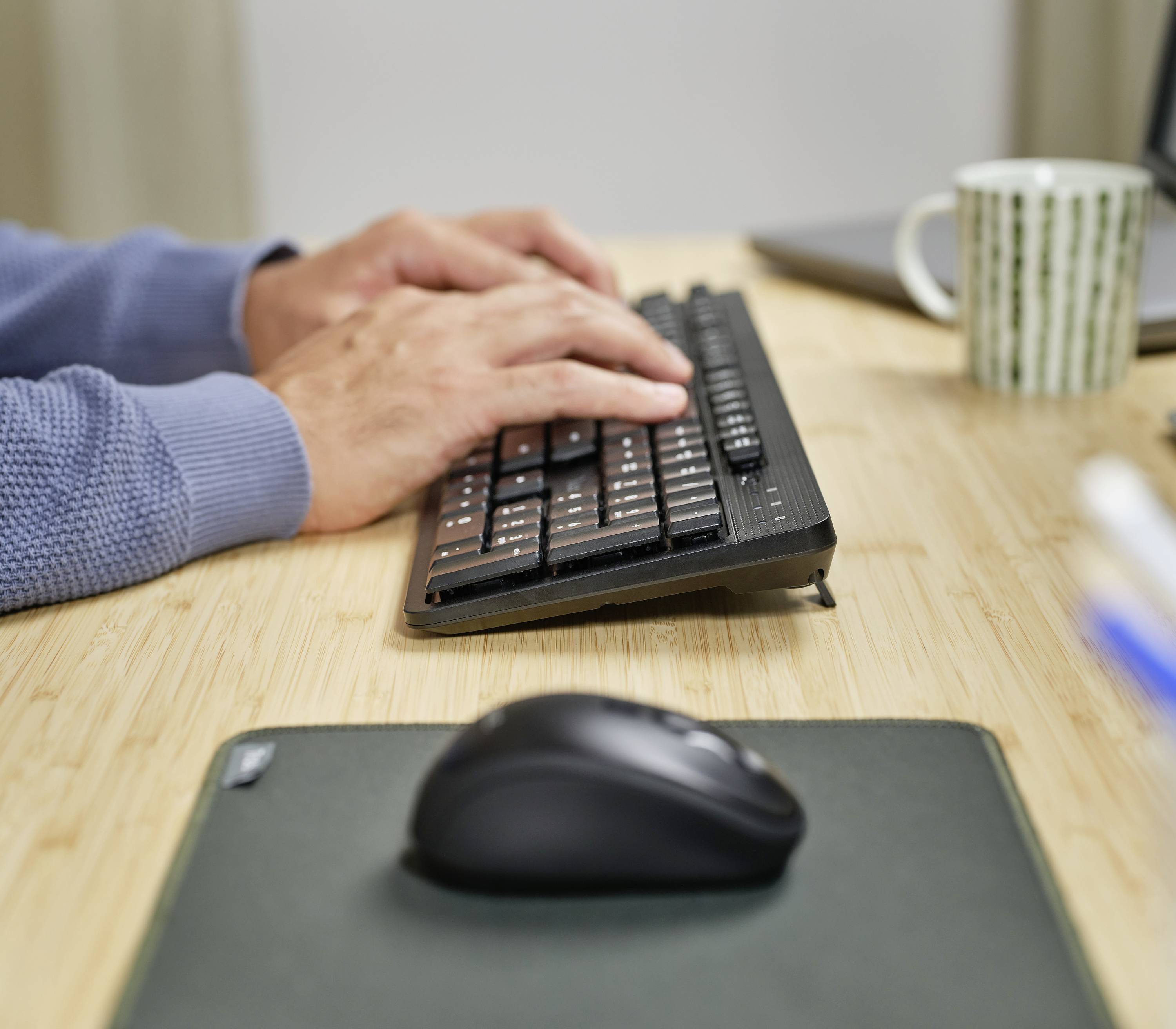 Hände tippen auf einer schwarzen Tastatur auf einem Schreibtisch. Eine Maus, ein Laptop und eine Tasse sind ebenfalls sichtbar.