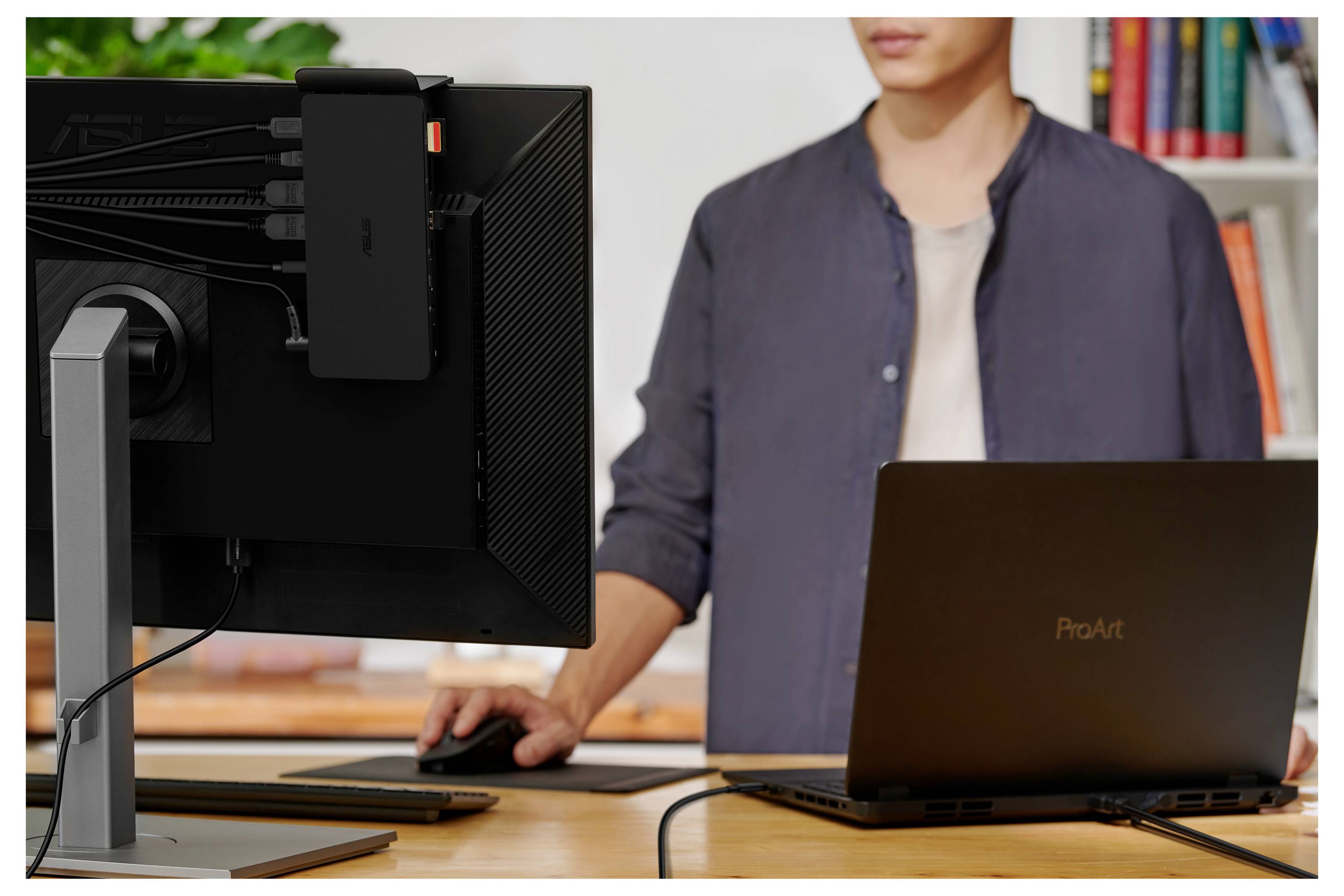 Eine Person steht vor einem Tisch mit einem Laptop und einem Monitor. Der Monitor zeigt keine spezifischen Inhalte. Im Hintergrund sind Bücherregale sichtbar.