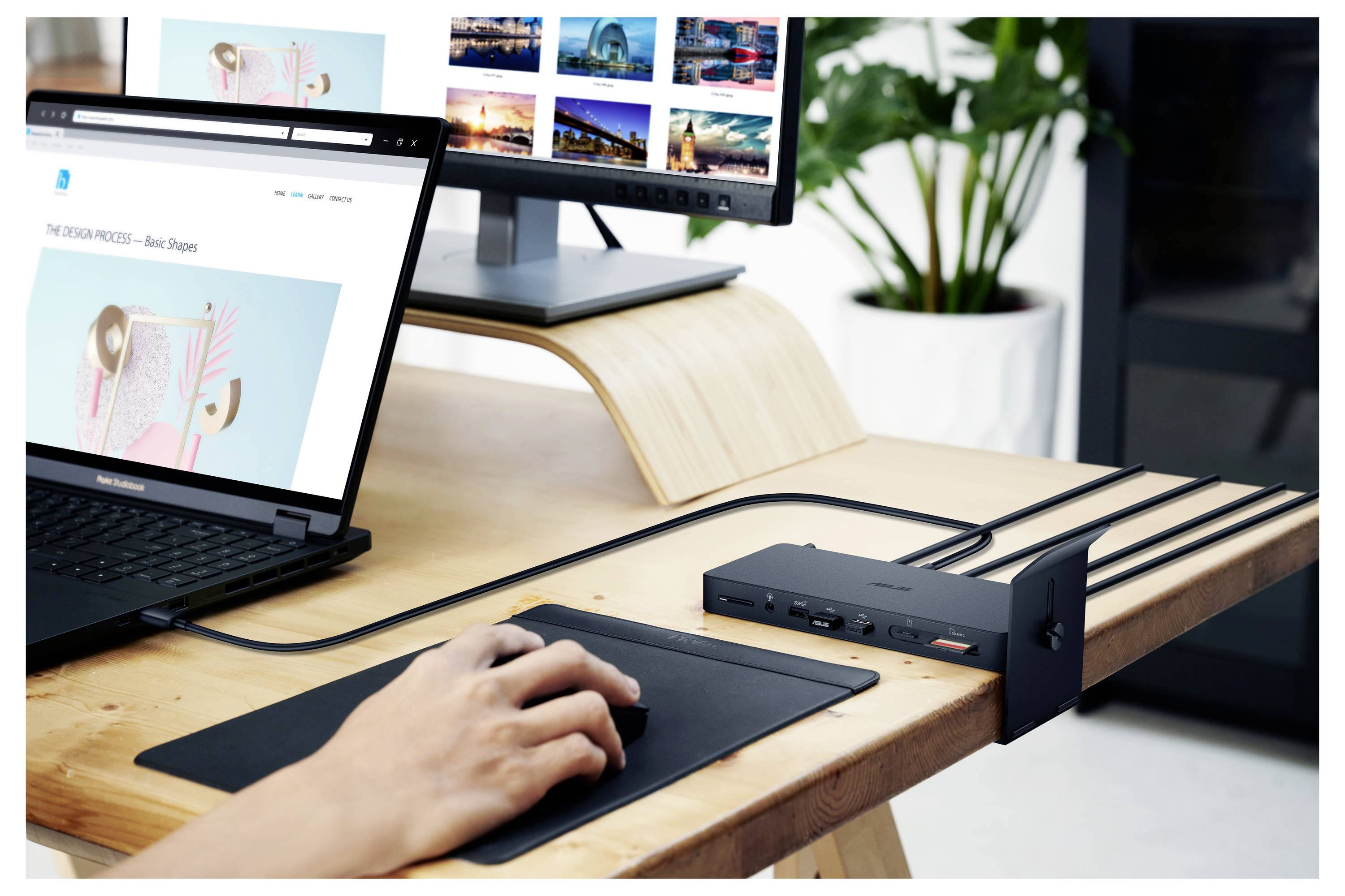Ein Schreibtisch mit einem Laptop, Monitor und Dockingstation. Eine Hand hält eine Maus. Pflanzen im Hintergrund.