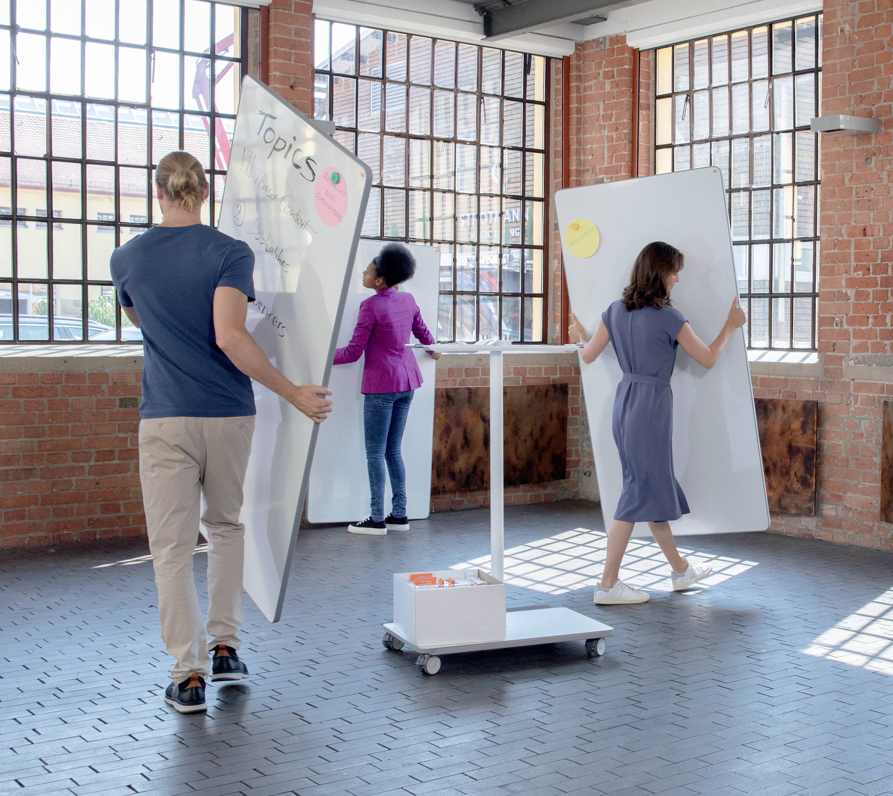 Drei Personen bewegen zwei große Whiteboards in einem hellen Raum mit Backsteinmauern und großen Fenstern, die in der Sonne leuchten.