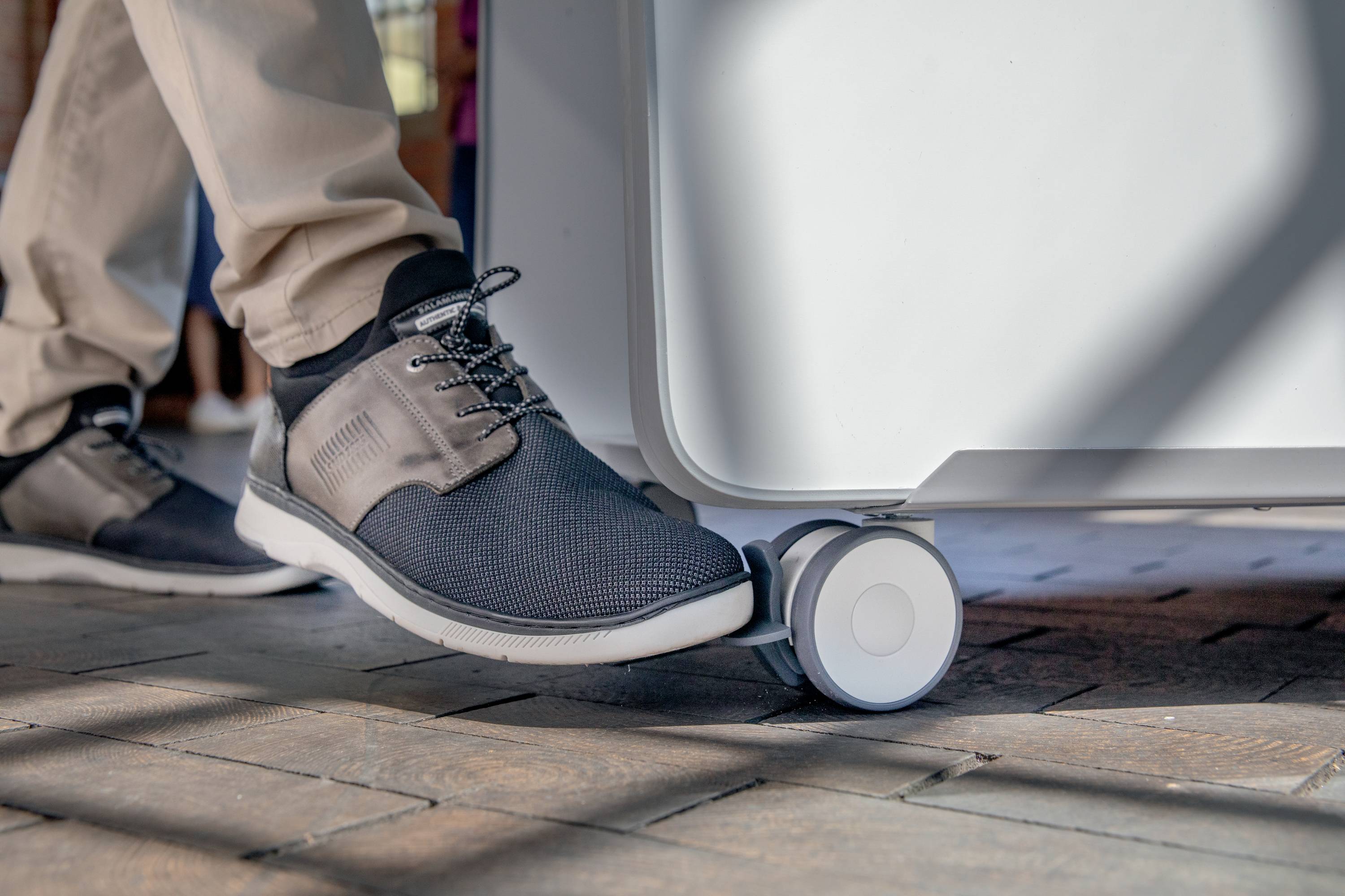 Ein Fuß, der auf einem runden Pedal eines kleinen Wagens oder Geräts steht, vermutlich um es zu bewegen. Trägt dunkelblaue Sneakers.