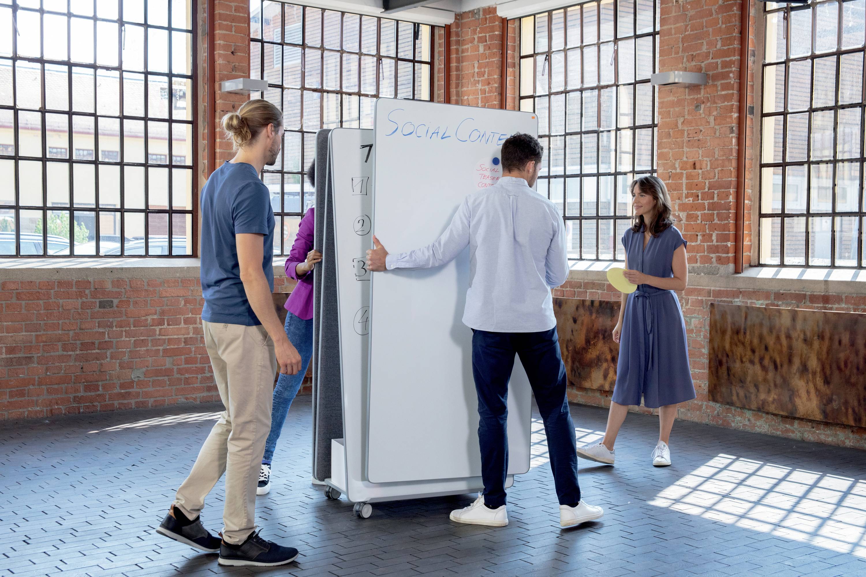 Eine Gruppe von fünf Personen überlegt vor einem weißen Board im Raum. Eine Person schreibt auf das Board. Reduzierte industrielle Umgebung.