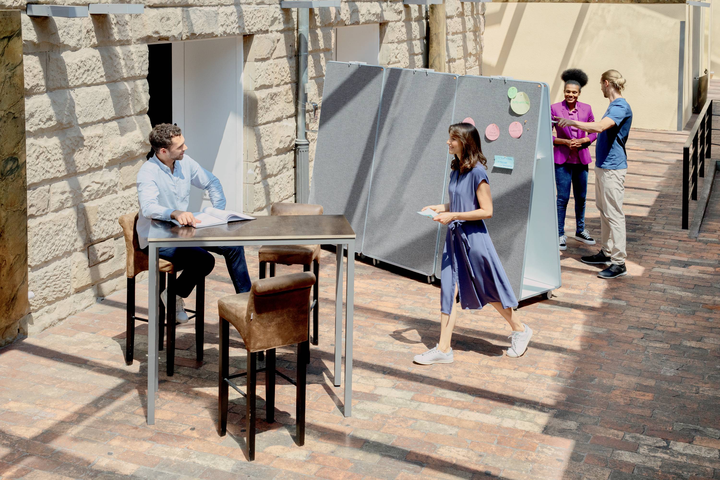 Personen arbeiten und diskutieren in einem offenen, sonnigen Innenhof. Eine Frau geht mit Notizen in der Hand. Im Hintergrund interagieren zwei Personen an einer Pinnwand.