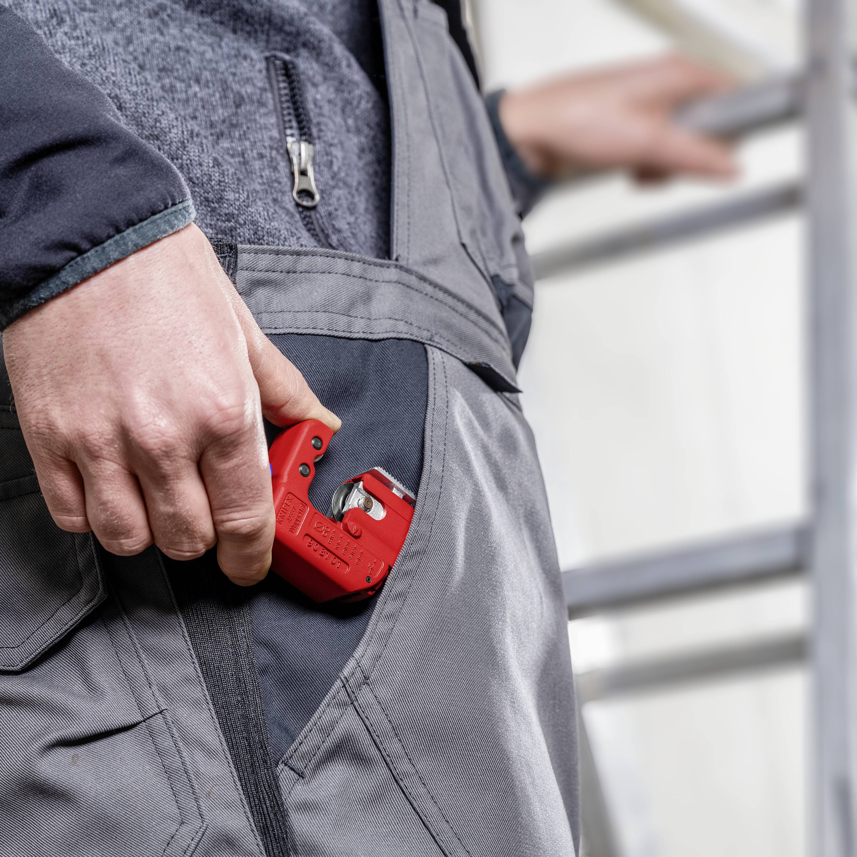 Eine Person steckt einen roten Rohrschneider in die Tasche eines grauen Arbeitsanzugs. Es handelt sich um eine Arbeitssituation.
