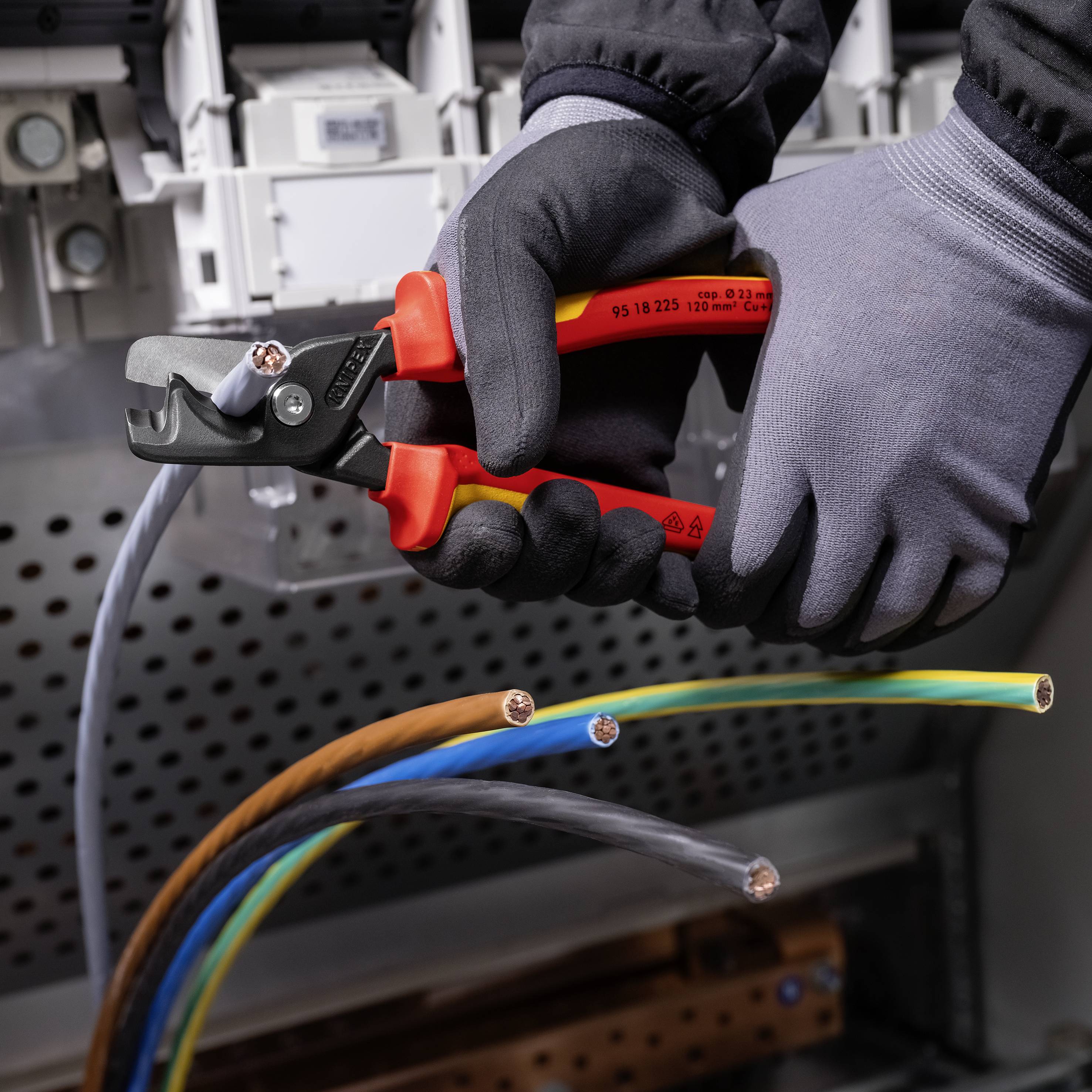 Eine Person mit grauen Arbeitshandschuhen schneidet mit einem roten Kabelschneider ein dickes Stromkabel vor einem Schaltschrank.