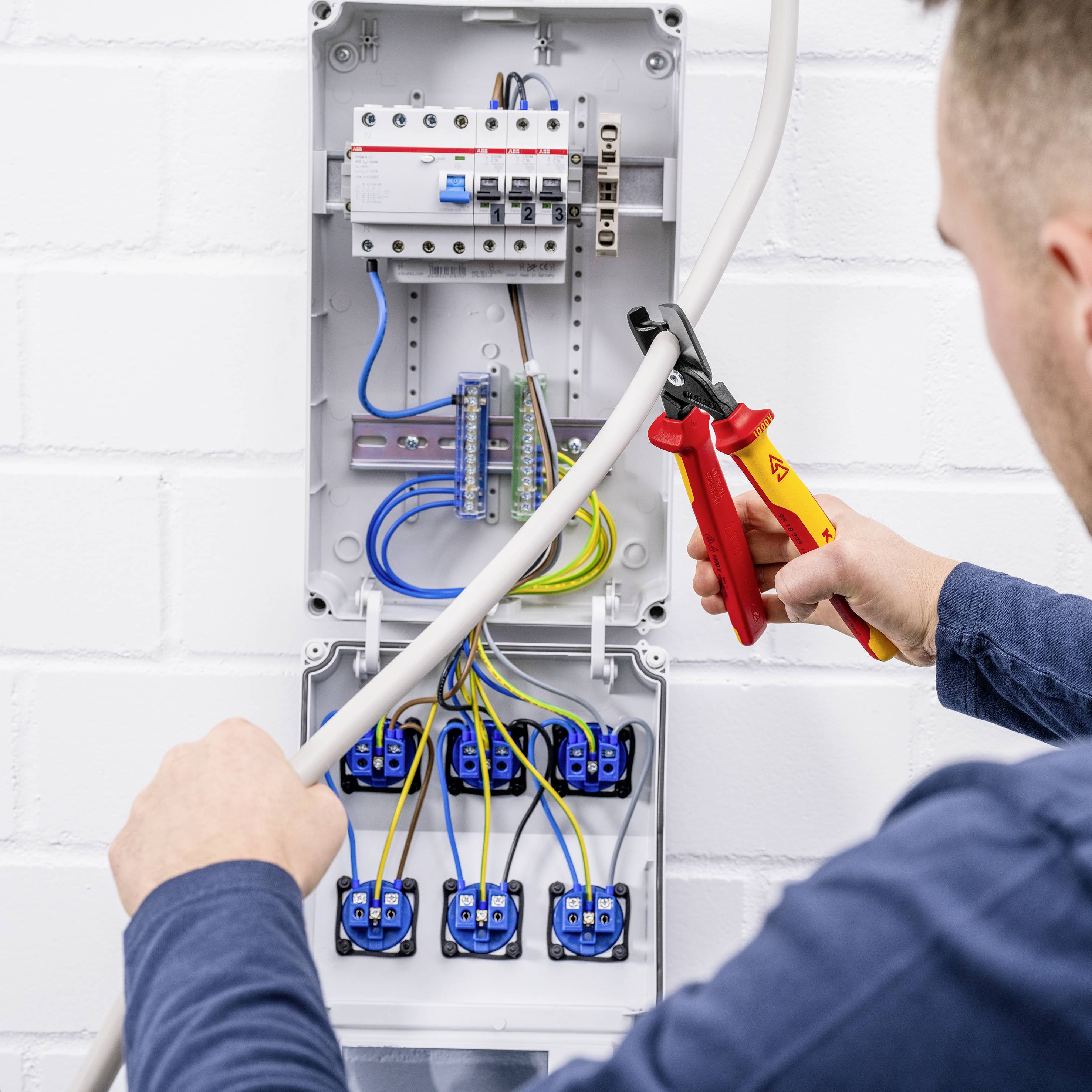 Eine Person arbeitet an einem geöffneten Stromverteilerkasten und verwendet ein rotes Werkzeug, um ein Kabel abzuisolieren.