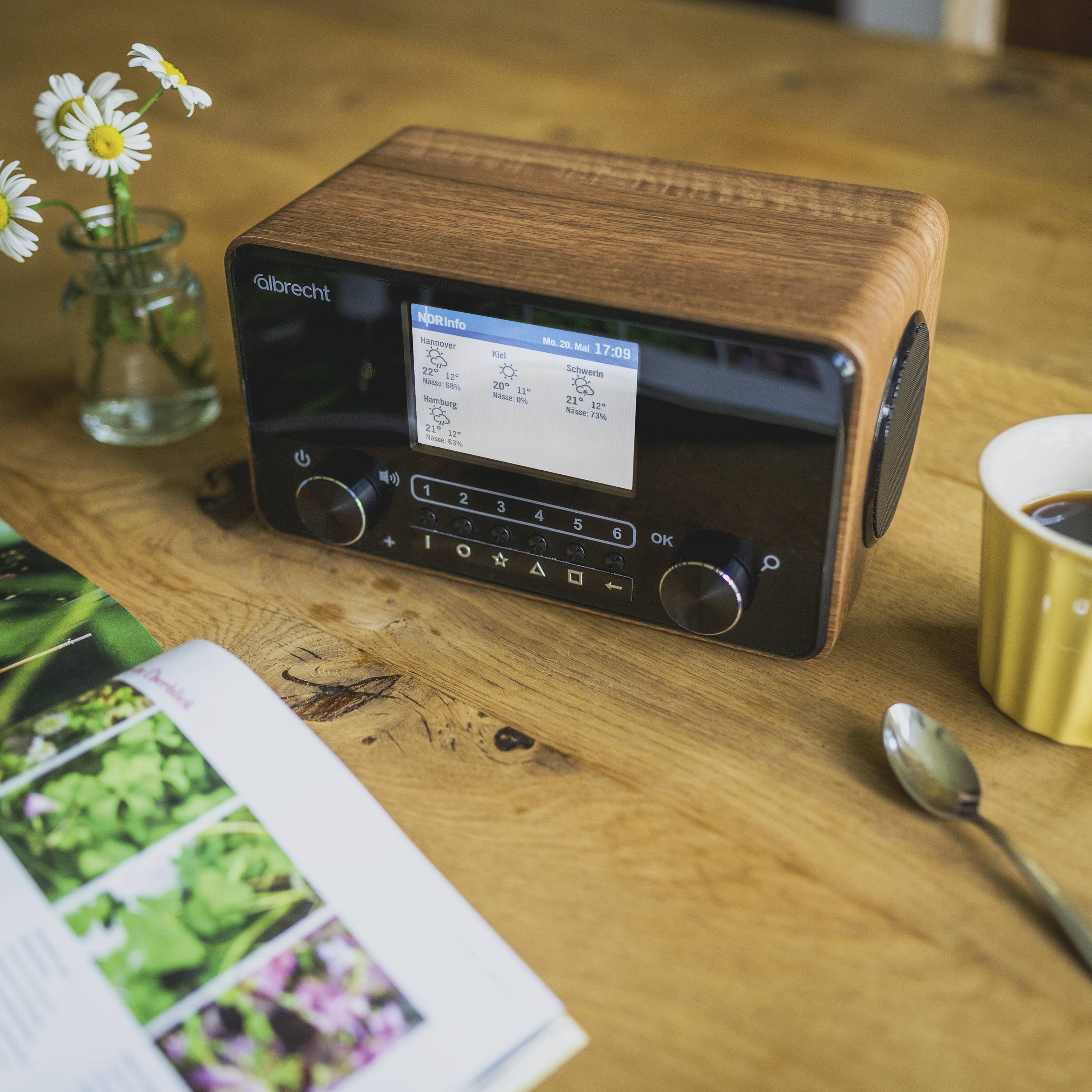 Retro-Radio auf Holztisch, umgeben von einer Blumenvase, einer Tasse Kaffee und einem Magazin, das aufgeschlagen ist.