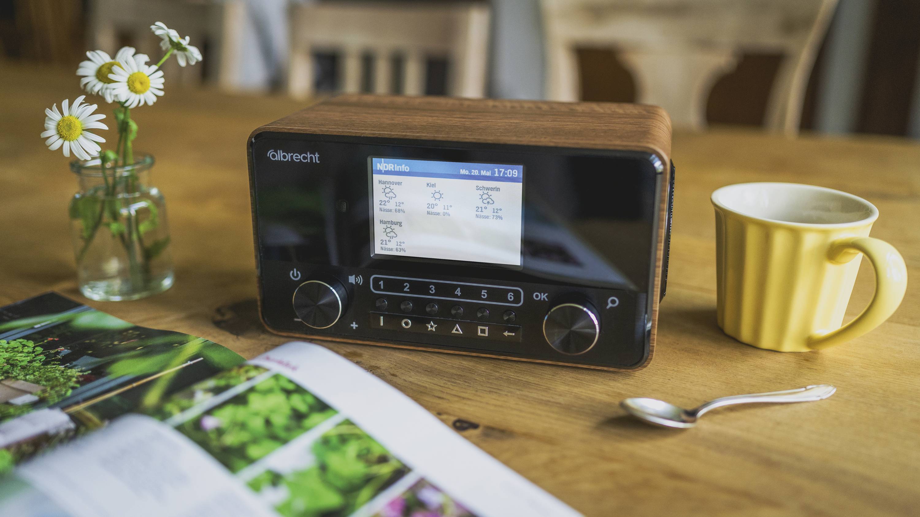 Altes Radio mit Holzdesign auf einem Tisch, daneben ein gelber Becher, ein Löffel, ein Magazin und ein Glas mit Gänseblümchen.