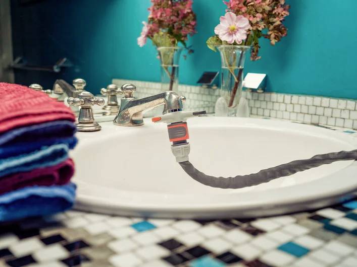 Ein Waschbecken mit fließendem Wasser, ein grauer Schlauch wird befüllt. Im Hintergrund stehen Blumenvasen, daneben gestapelte Handtücher.