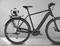 Schwarzes E-Bike mit gepolstertem Sitz und Ballhalterung am Hinterradträger, darin ein Fußball. Steht vor grauem Hintergrund.