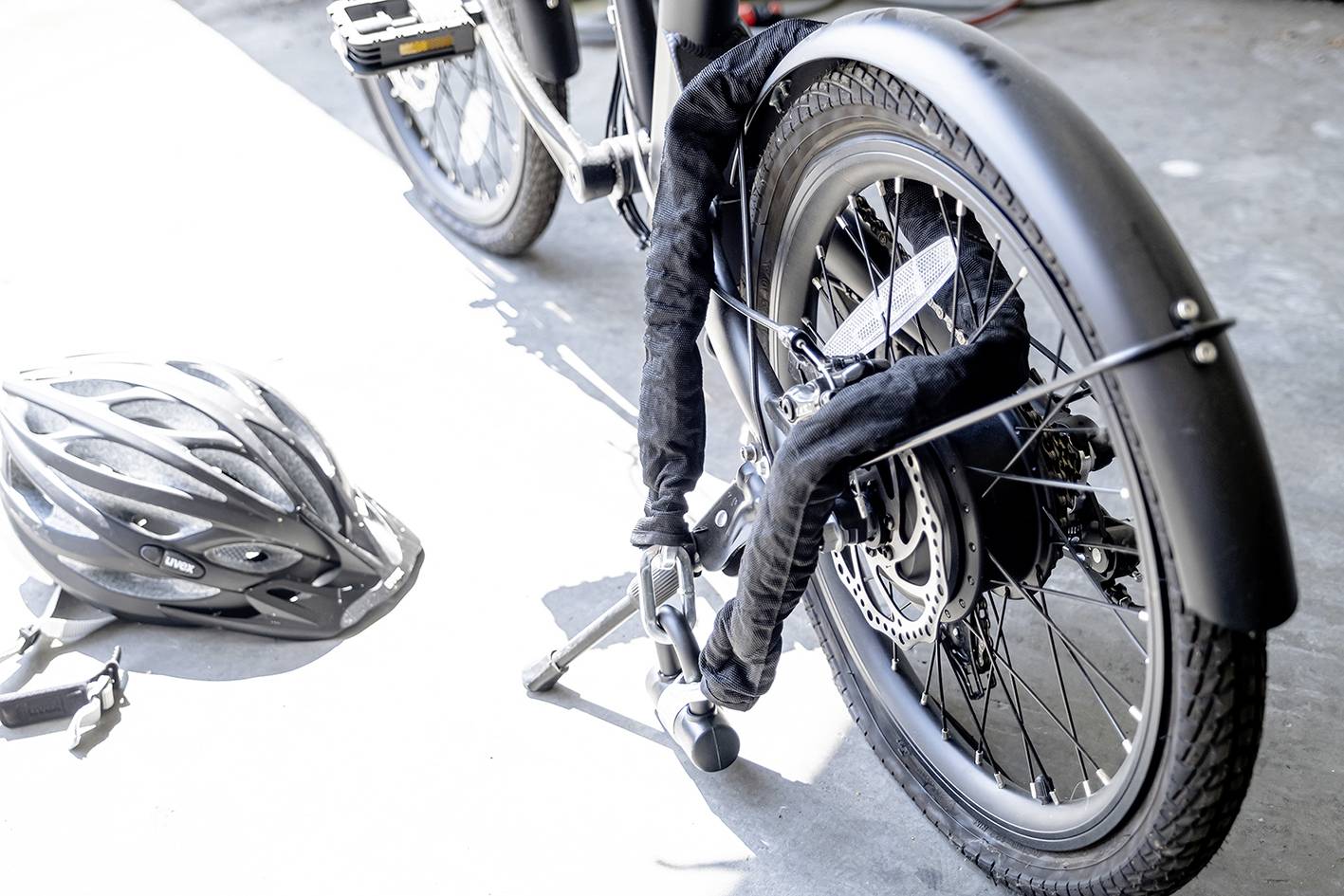 Ein schwarzes Fahrrad mit dickem Schloss am Hinterrad steht auf Beton. Ein schwarzer Fahrradhelm liegt daneben auf dem Boden.