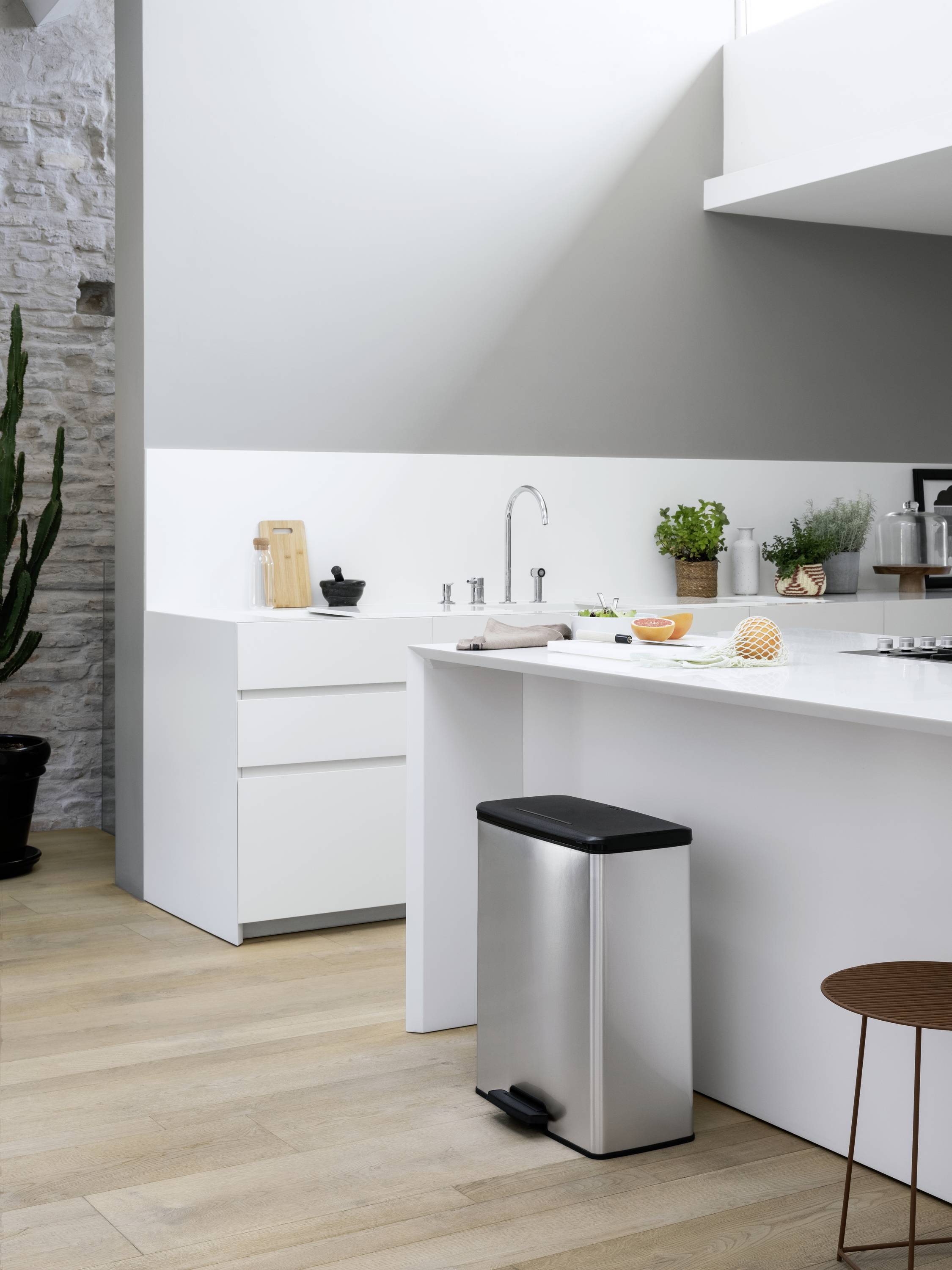 Moderne Küche mit weißem Interieur und Edelstahl-Mülleimer im Vordergrund. Helle Arbeitsplatten mit Kräutern und Küchenutensilien.