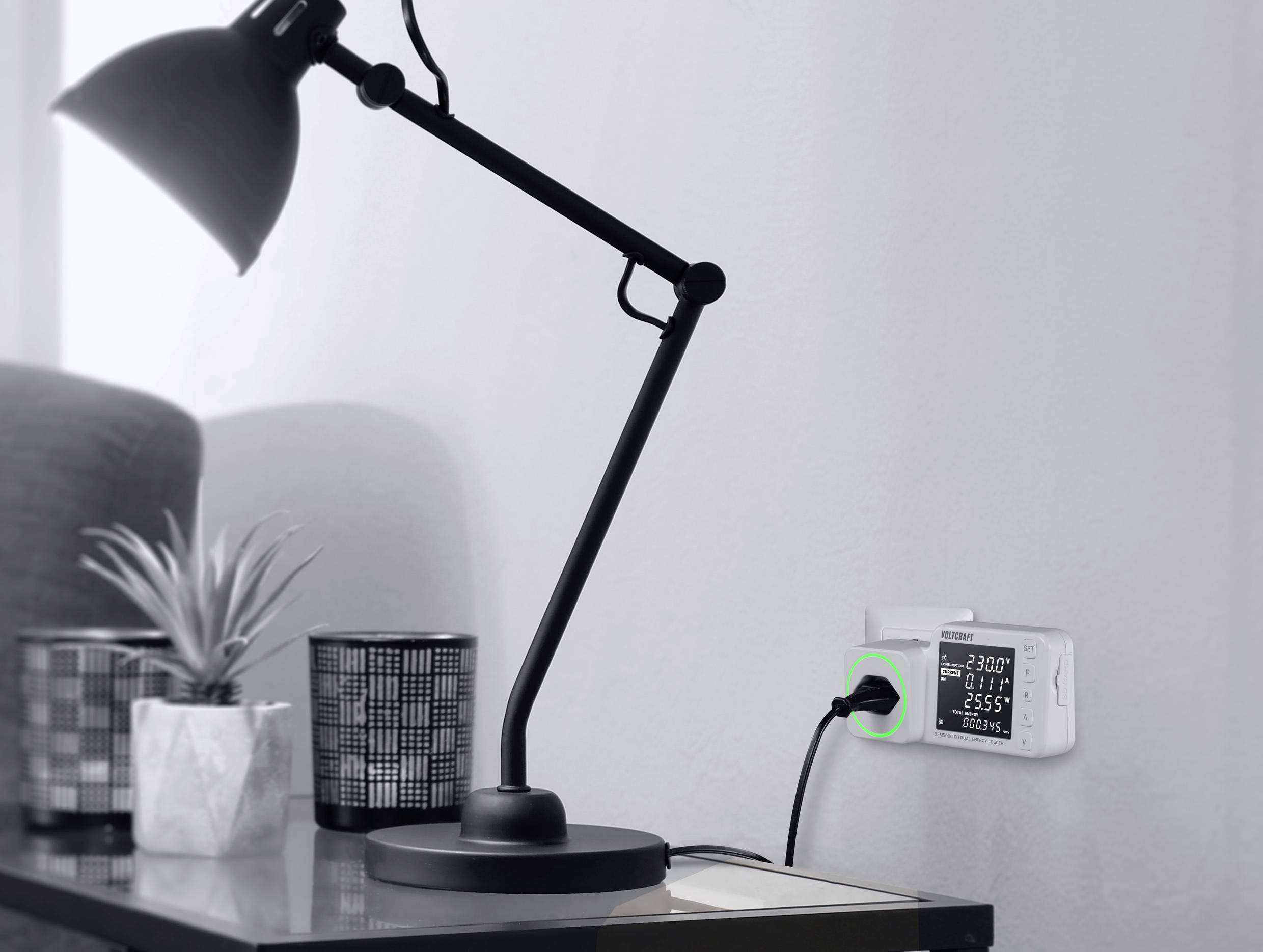 Eine Schreibtischlampe neben einer Vase und einem Becher. Ein Überwachungsgerät steckt in der Wand, zeigt Werte wie Spannung an.