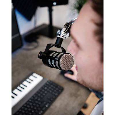 Ein Mensch spricht in ein Mikrofon in einem Studio. Im Vordergrund sind eine Computertastatur und ein MIDI-Keyboard zu sehen.