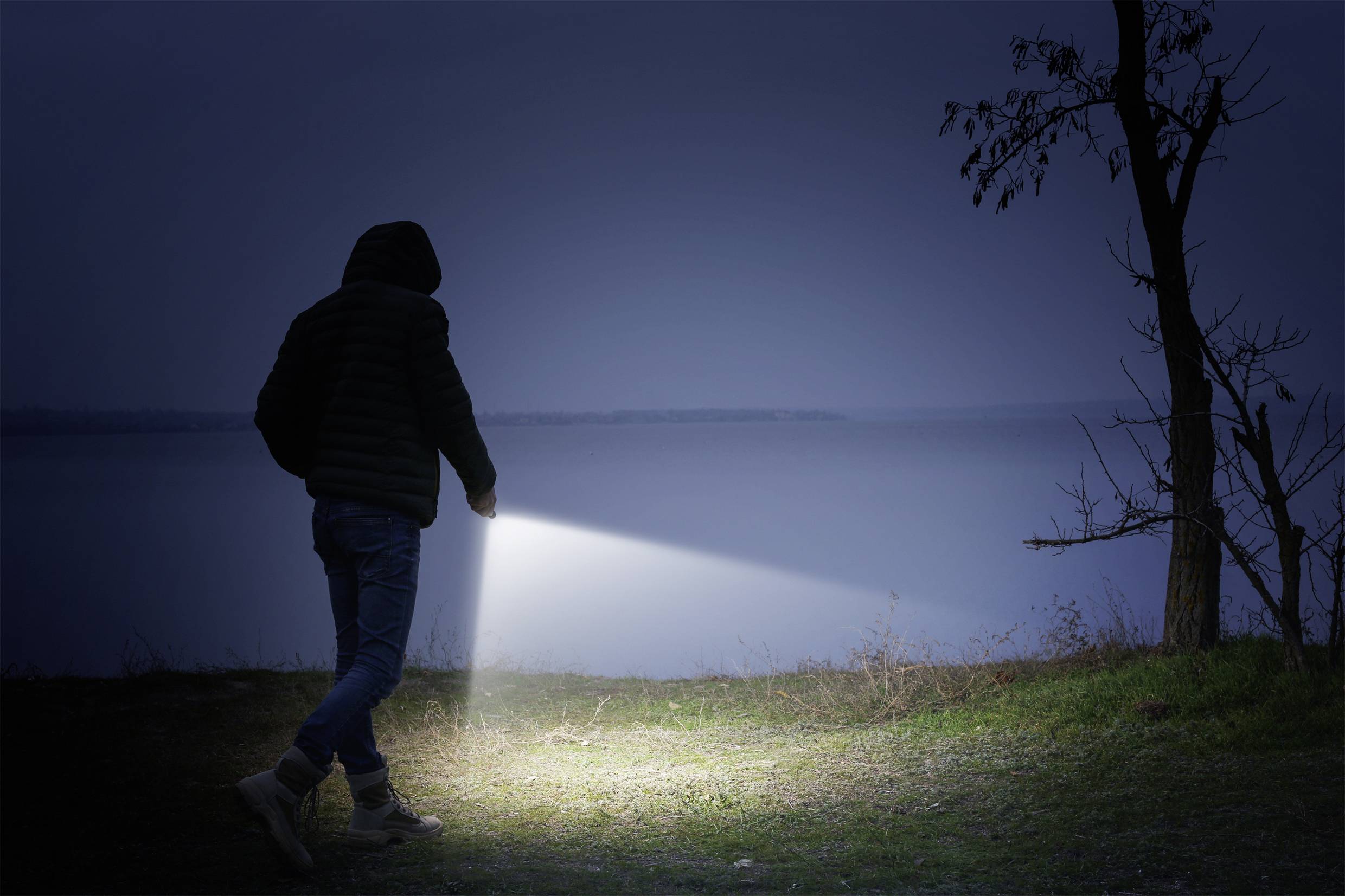 Person mit Taschenlampe geht nachts am Ufer entlang, beleuchtet den Boden. Atmosphäre ist dunkel und ruhig mit wenig sichtbaren Details.