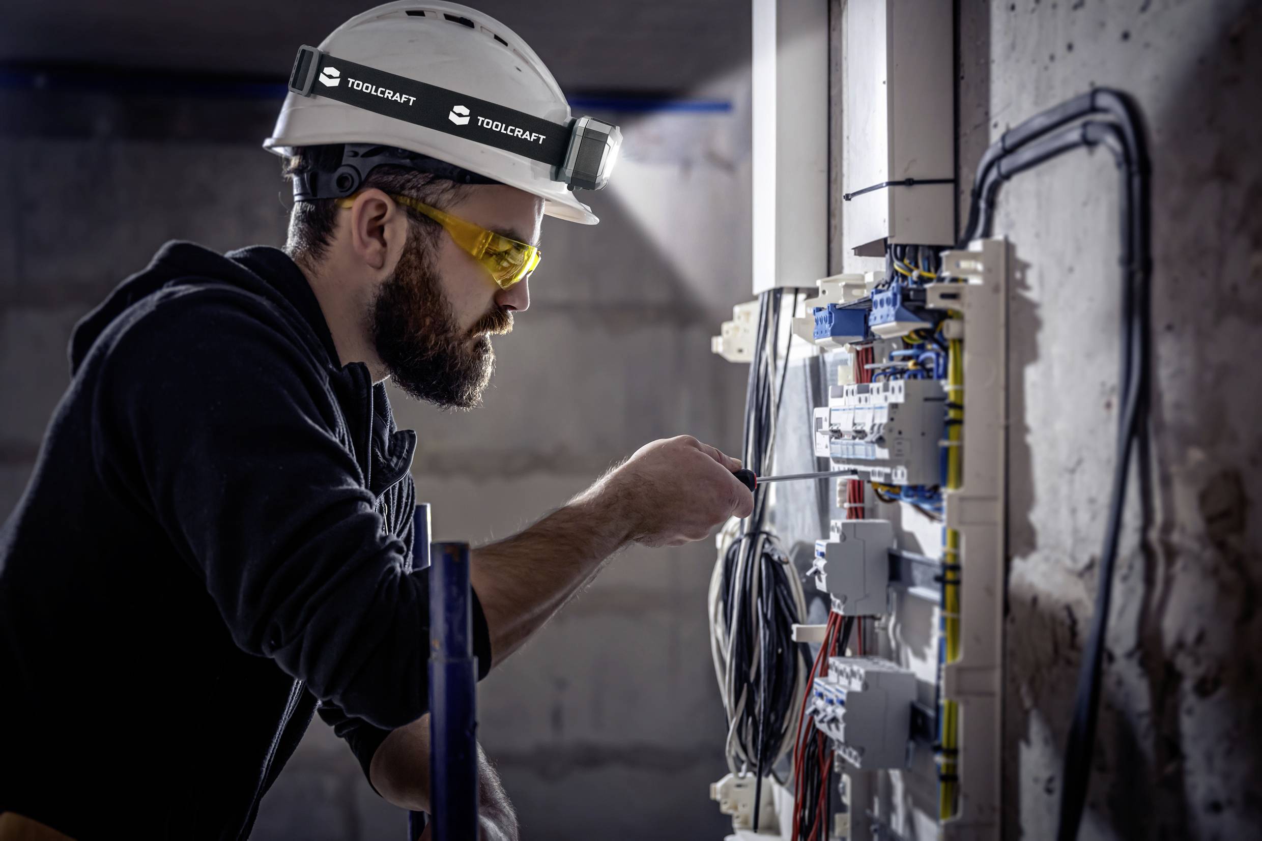 Ein Elektriker mit Schutzhelm und Schutzbrille arbeitet an einem offenen Schaltschrank, prüft und justiert die elektrischen Anschlüsse.