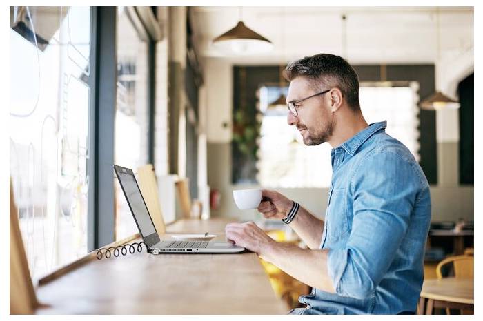 Ein Mann sitzt in einem Café am Laptop und hält eine Tasse Kaffee. Er konzentriert sich auf den Bildschirm.