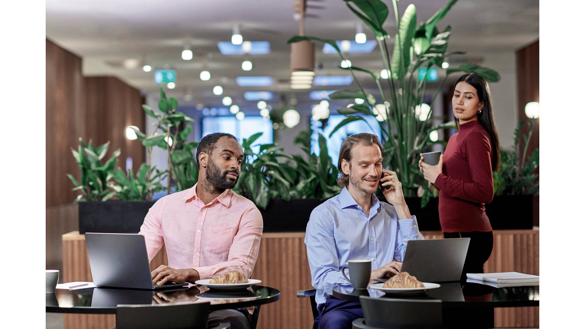 Drei Personen in einem modernen Café mit Laptops und Getränken. Zwei sitzen und arbeiten an Laptops, die dritte hält eine Tasse.