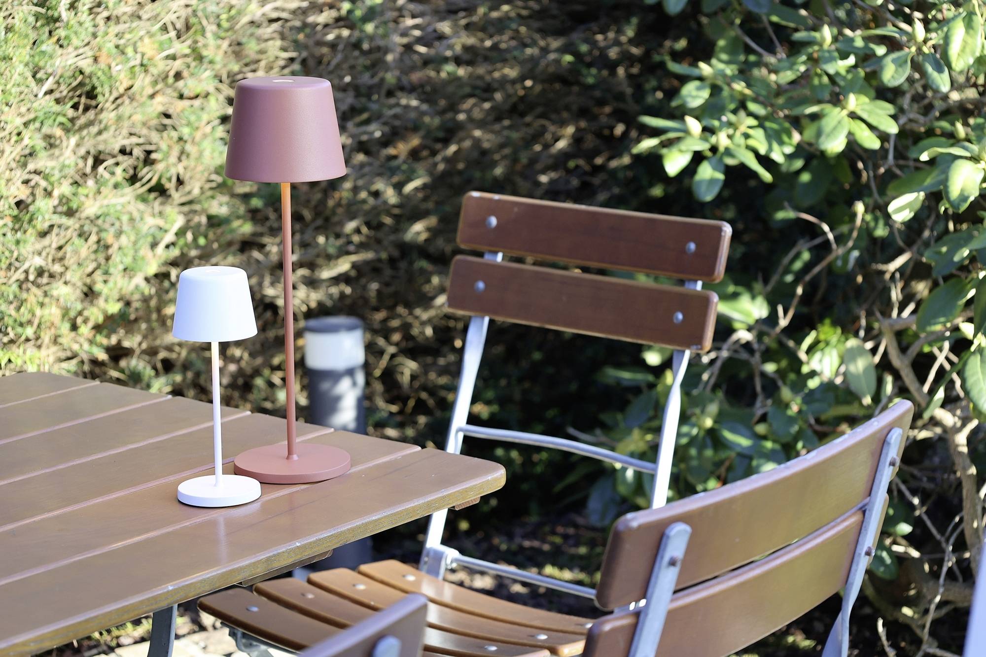 Ein Holz-Bistrotisch mit zwei Stühlen steht im Freien. Auf dem Tisch sind zwei Lampen, eine rot, eine weiß. Im Hintergrund sind Pflanzen.