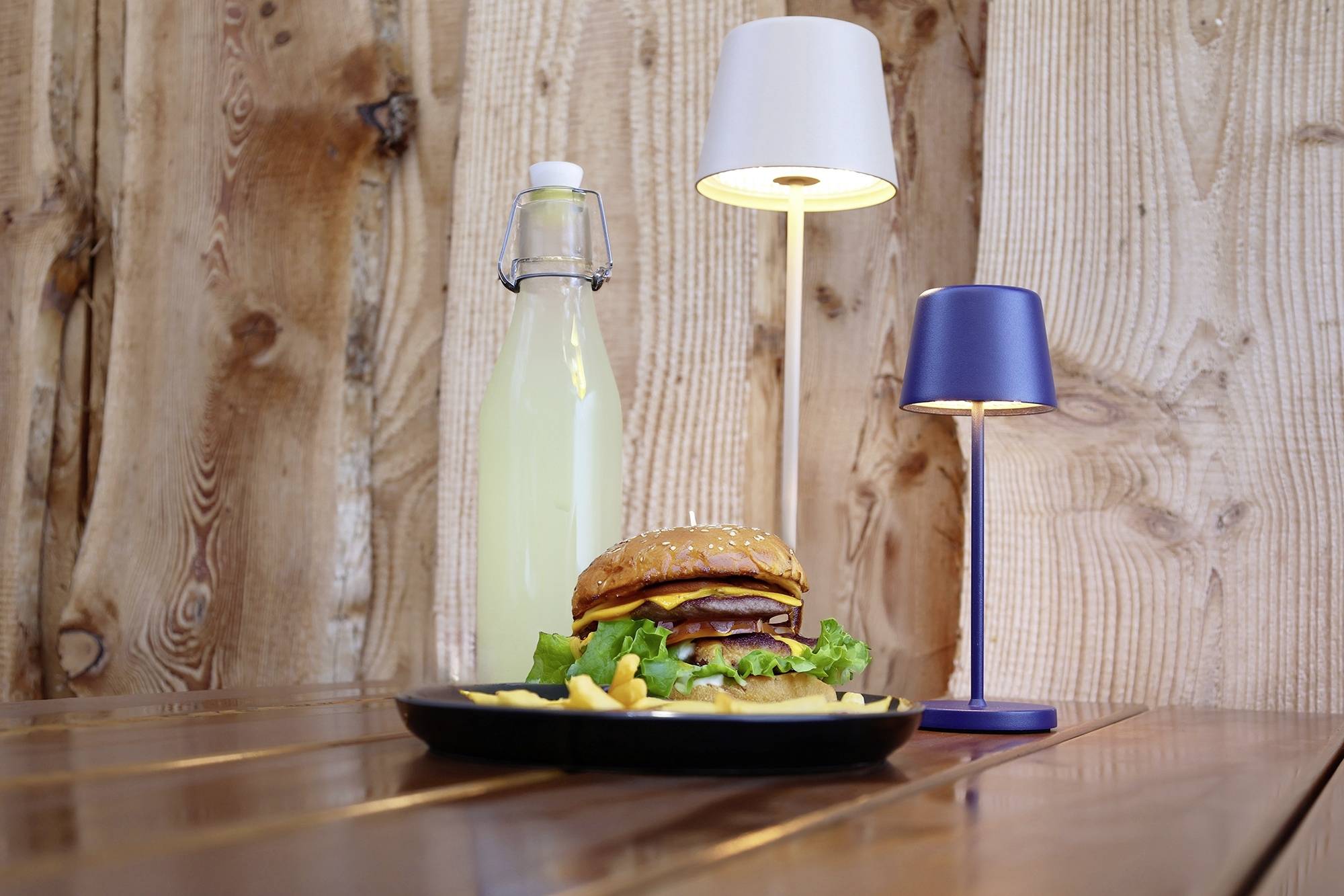 Ein Teller mit einem Burger und Pommes steht auf einem Holztisch. Daneben eine Flasche Limonade und zwei kleine Tischlampen.