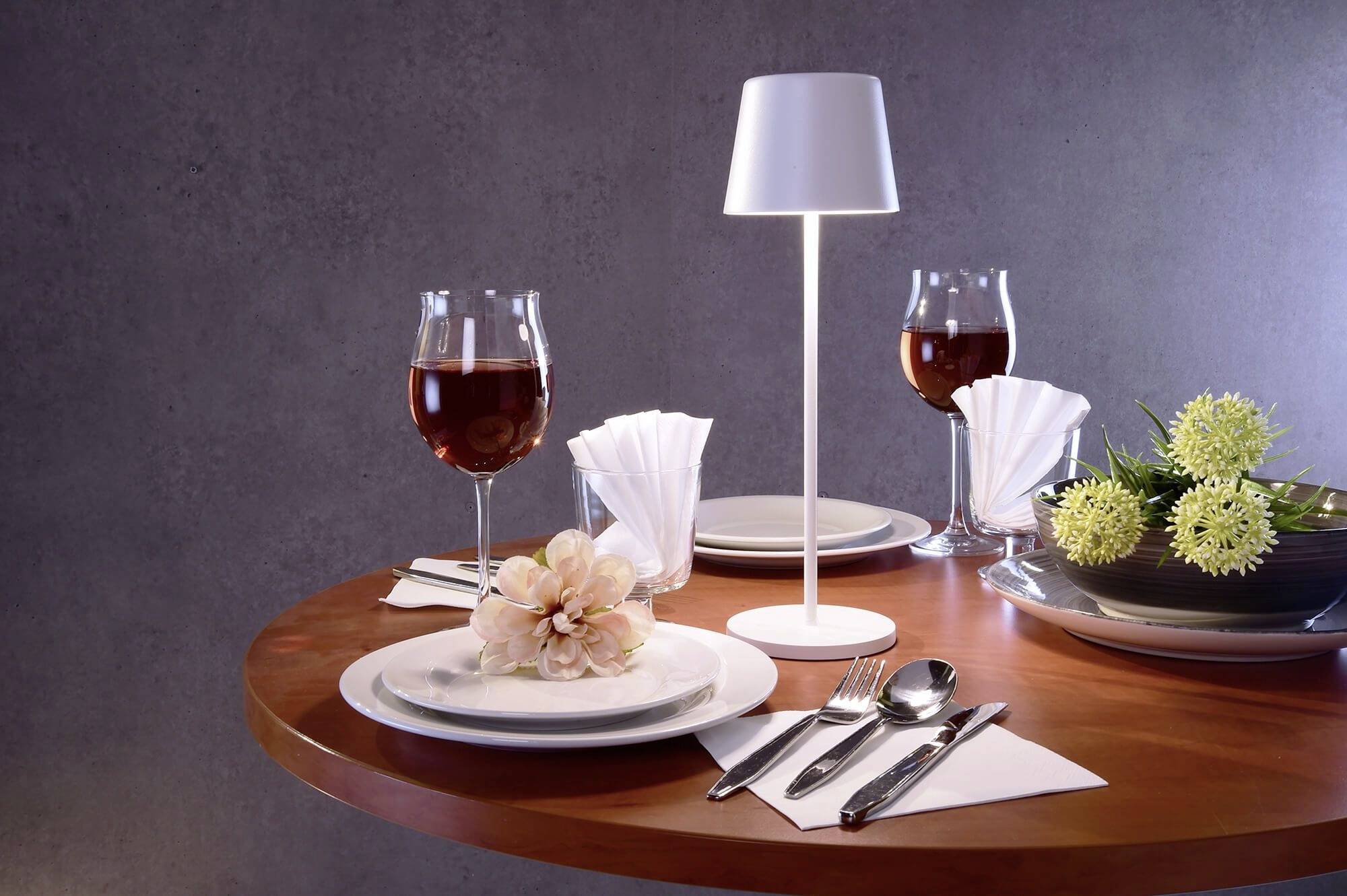Ein elegant gedeckter Tisch mit zwei Weingläsern, einem Blumenarrangement, weißer Tischlampe und dekorativen Servietten.