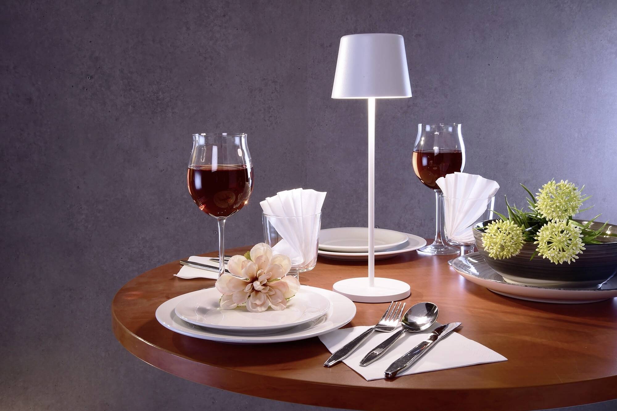 Ein runder Holztisch ist festlich gedeckt mit zwei Weingläsern, einer weißen Tischlampe, Besteck, Papierservietten und Blumenarrangement.