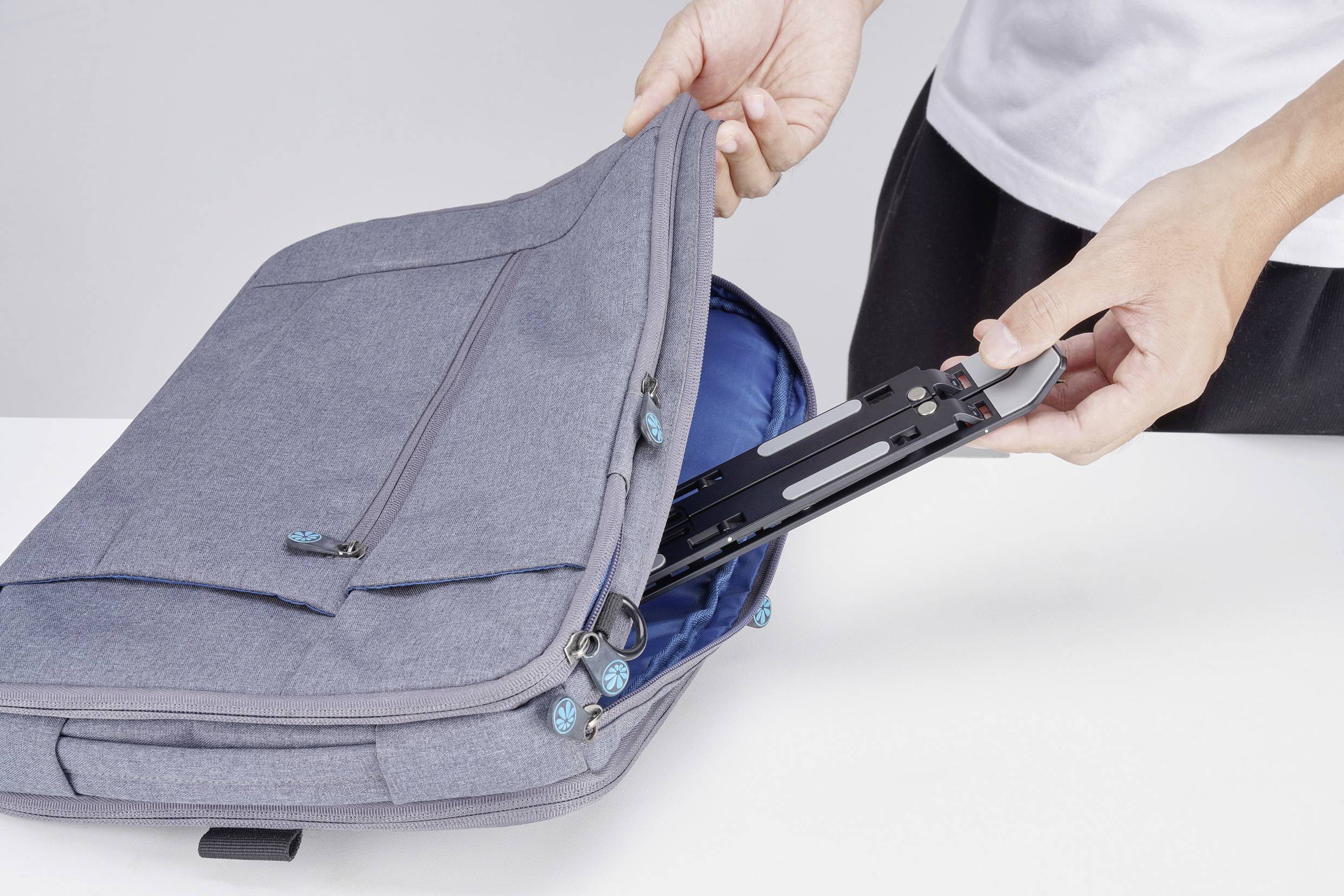 Eine Person stellt einen schwarzen Laptop-Ständer zum Zusammenklappen in eine graue Aktentasche auf einem weißen Tisch und bereitet sich auf Reise oder Arbeit vor.