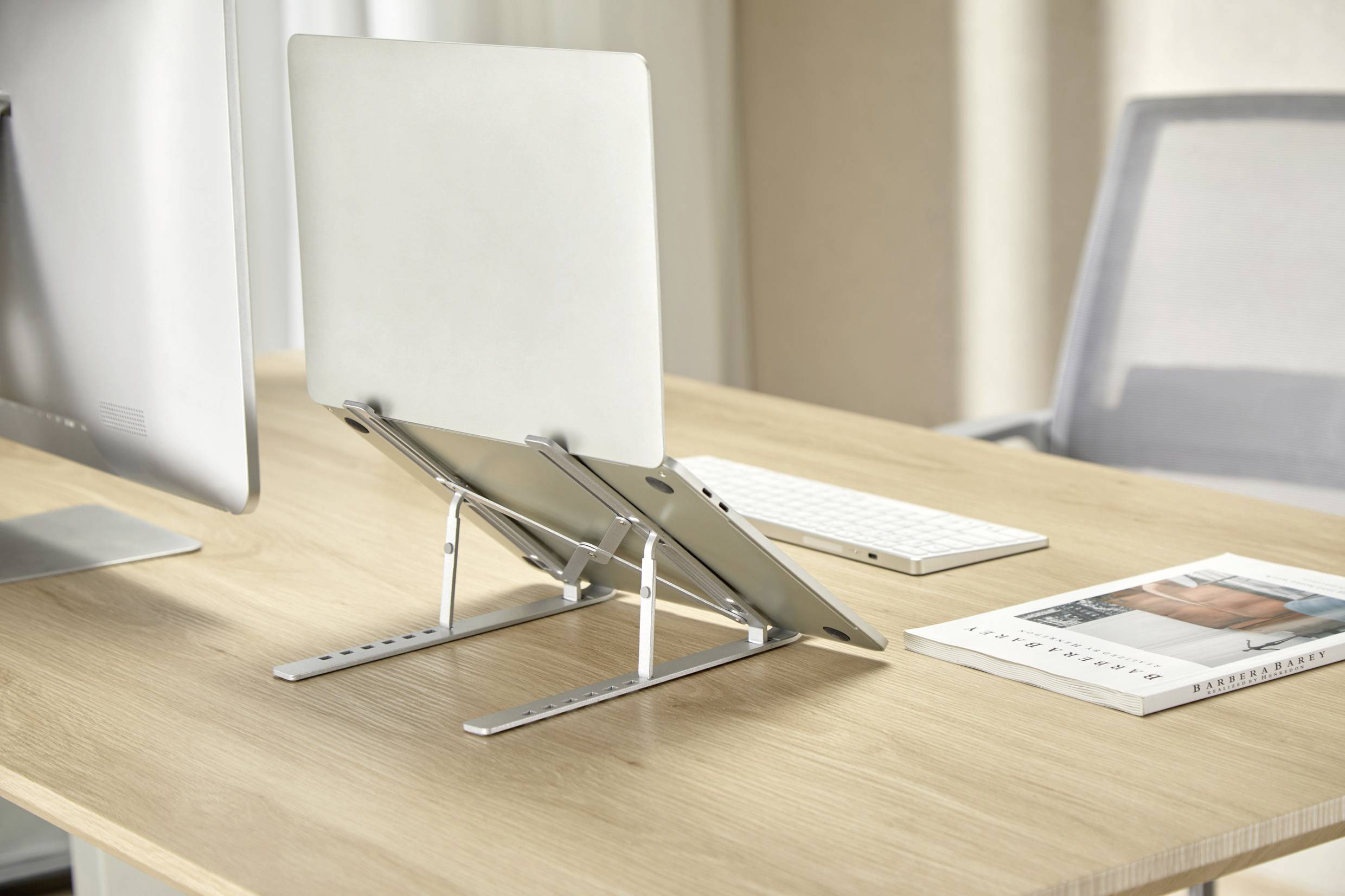 Ein Tablet und ein Laptop, beide auf verstellbaren Metallständern, stehen auf einem Holzschreibtisch neben einem Monitor, einer Tastatur und einer Zeitschrift.