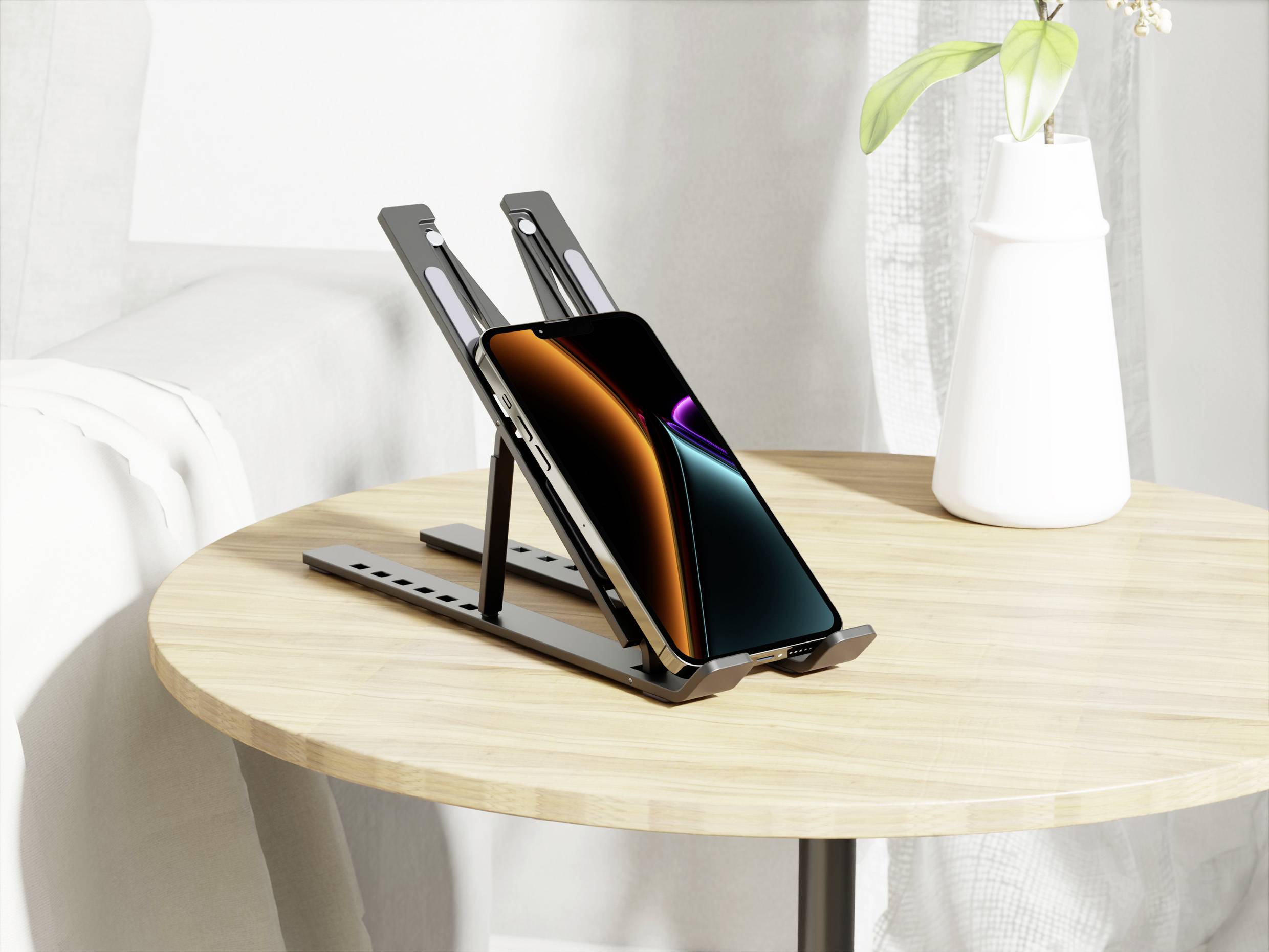 Ein Smartphone mit einem bunten Bildschirm liegt auf einem verstellbaren schwarzen Ständer, der auf einem hellbraunen Holztisch neben einer weißen Vase platziert ist.